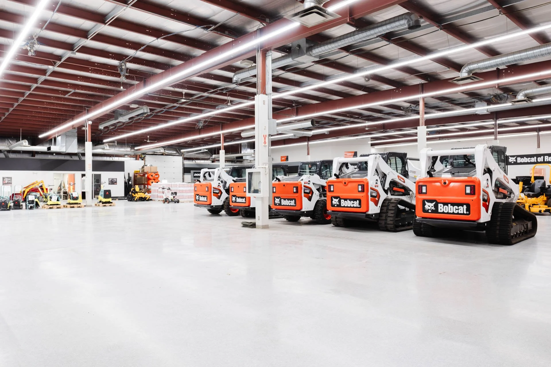 Indoor warehouse with new Bobcat compact track loaders parked in a row, high ceiling with exposed pipes, bright lighting, and additional construction equipment on the left.
