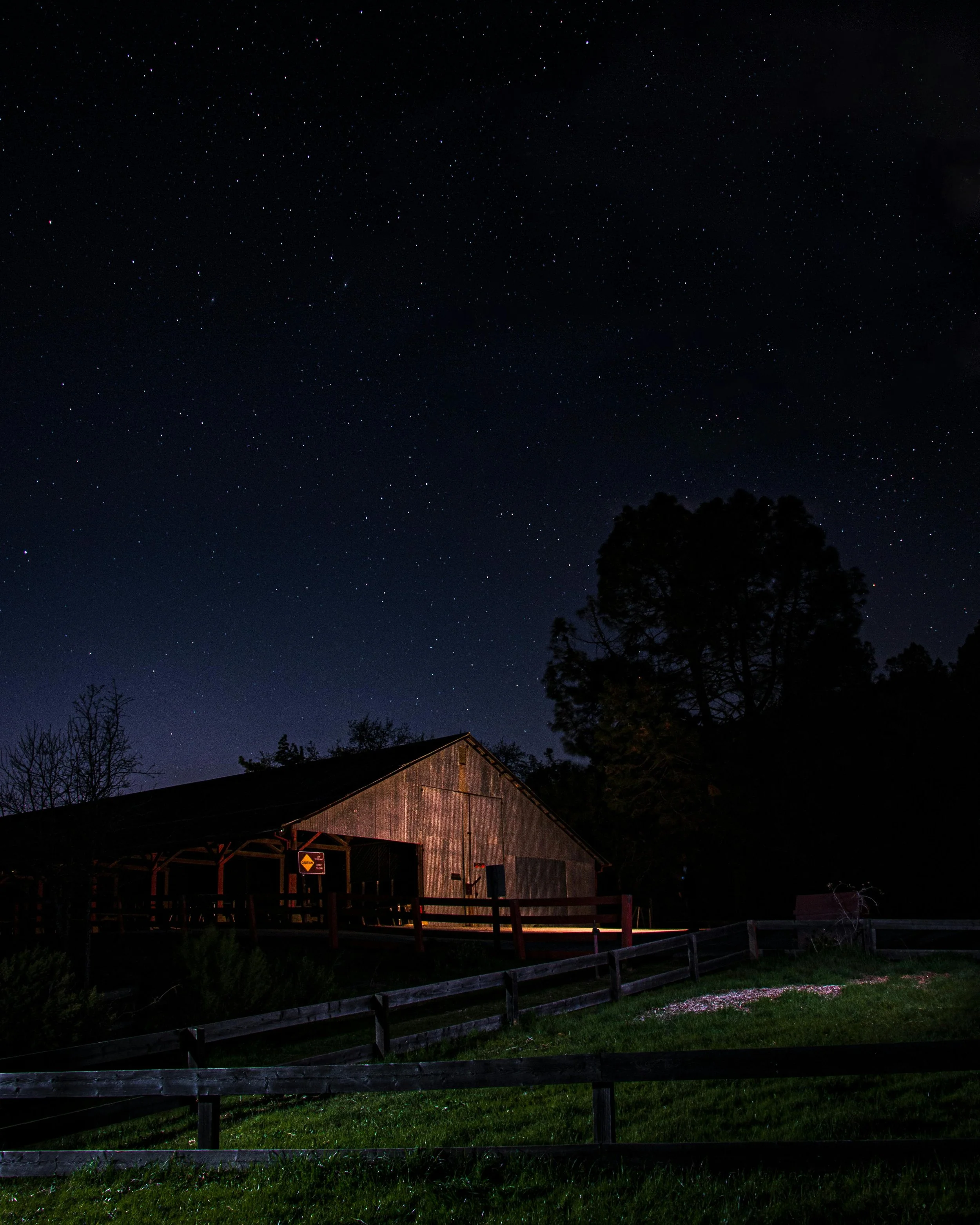 Nighttime scene of a rustic barn with a slanted roof, illuminated slightly, surrounded by trees and a grassy area with a wooden fence, under a sky filled with numerous visible stars.