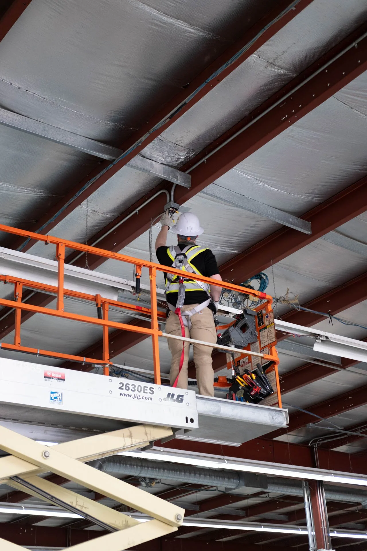 A construction worker wearing a white safety helmet, harness, and safety gear is working on electrical installation from an orange scissor lift inside a building with exposed ceiling beams, pipes, and wiring.