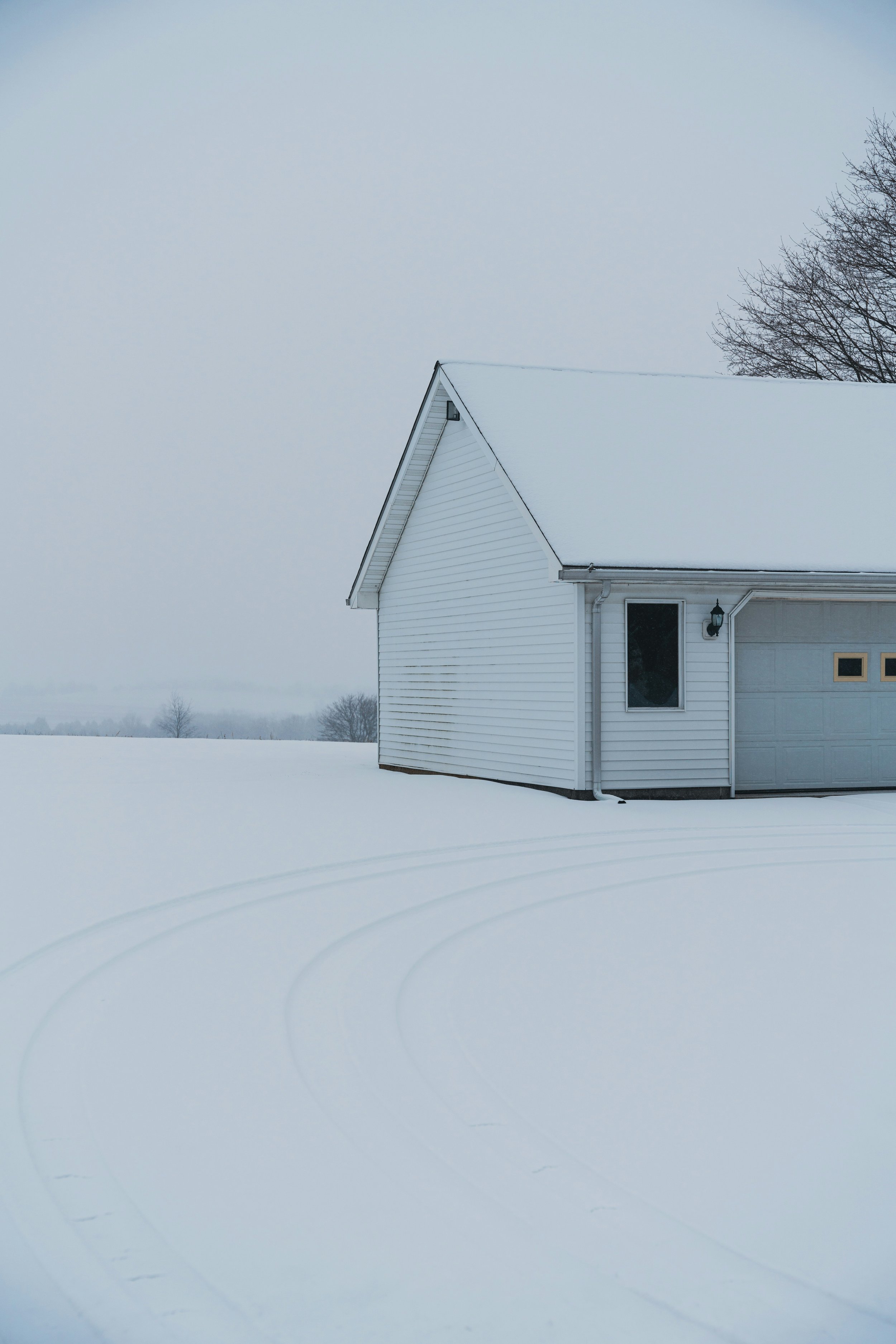 A white house with a snow-covered roof, a small window, and a garage door, set against a snowy landscape with tire tracks and a gray overcast sky.