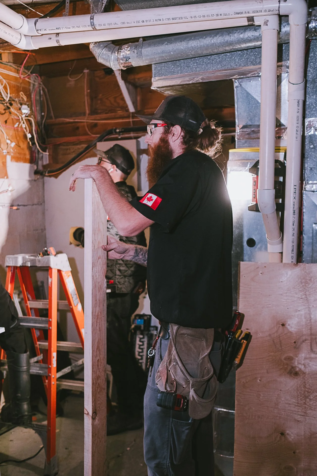 A man with a long beard, glasses, and a black baseball cap is working in a construction or renovation setting, holding a wooden plank. He has a glove in his left hand and is wearing a black shirt with a Canadian flag patch on the sleeve. He has a tool belt with various tools and a cordless drill on his hip. In the background, another person wearing a cap and gloves is working, and there is an orange ladder, exposed pipes, and wooden panels.