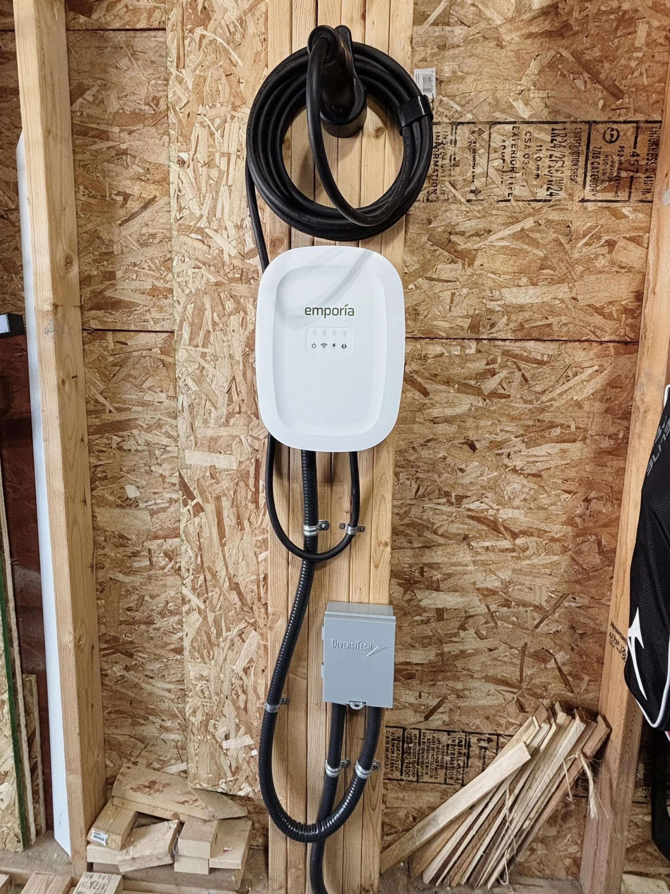 An electric vehicle charging station mounted on plywood wall with a connected black charging cable, a white control unit labeled 'Emporia,' and a gray box labeled 'Diverse Tech' at the bottom, surrounded by construction materials.