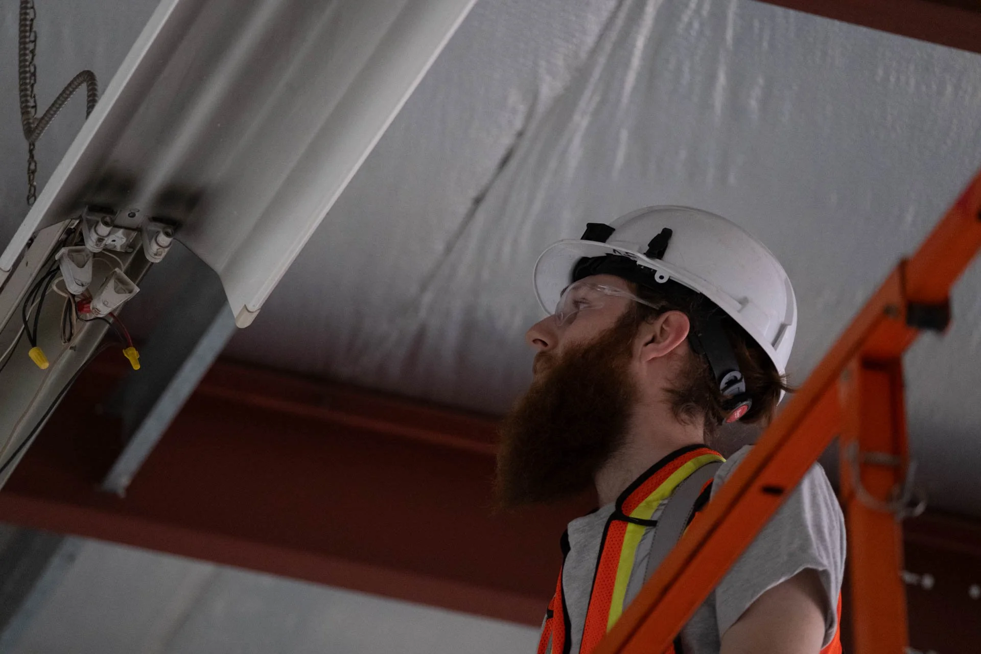 A technician working on electrical wiring on a ceiling, standing on an orange ladder, wearing a white hard hat, safety goggles, and a high-visibility safety vest.