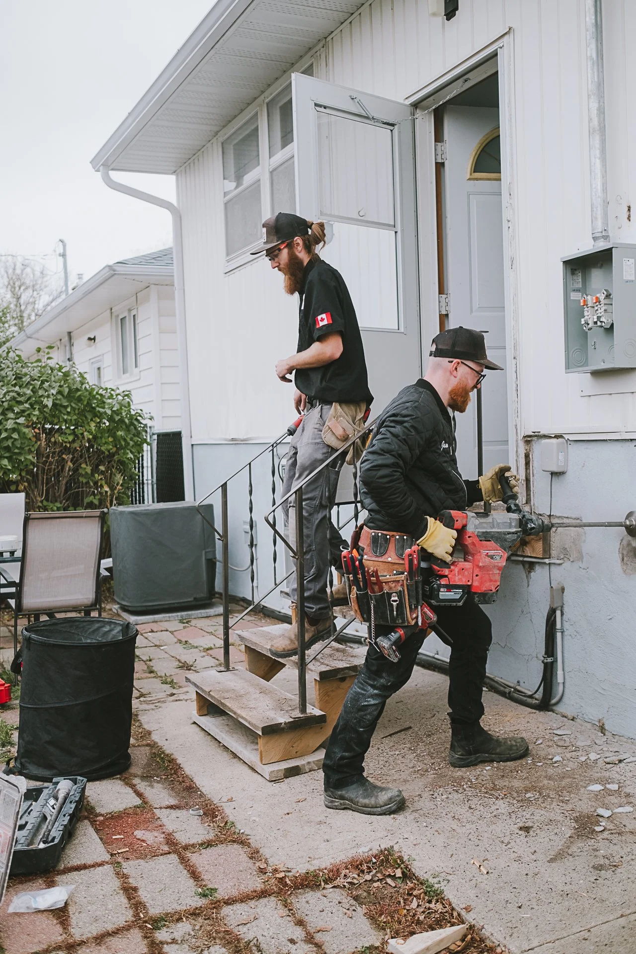 Two workers repairing the exterior wall of a house, one standing on stairs with a tool, and the other using a power drill on the wall.