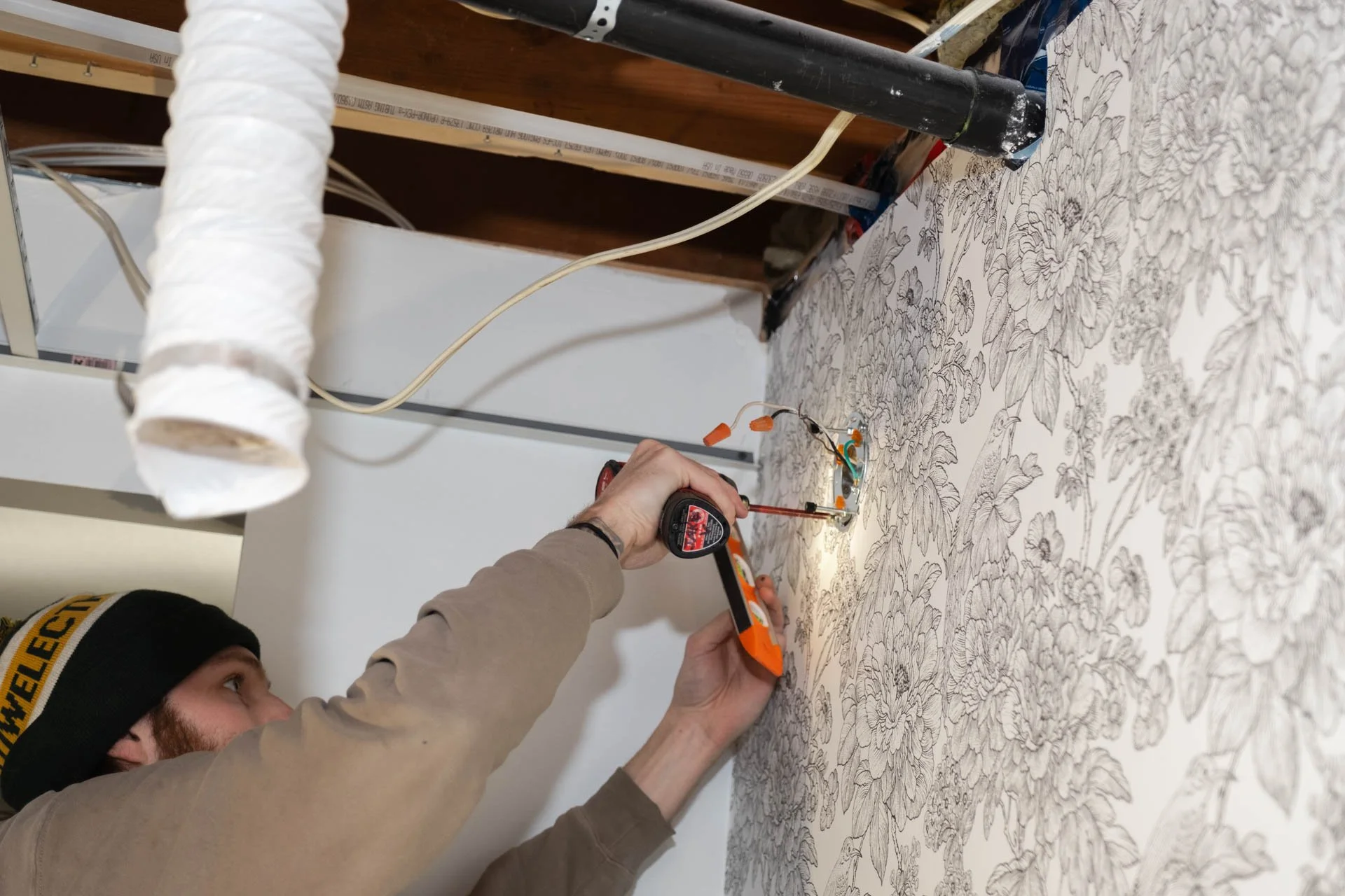 Person testing electrical wiring with a screwdriver and measuring tape on a floral wallpapered wall.