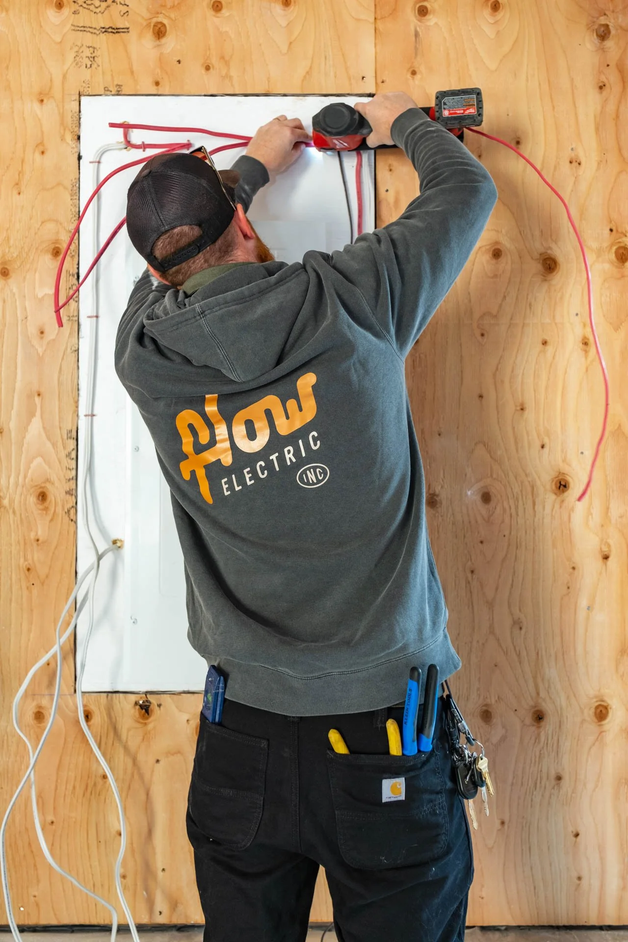 Man installing electrical wiring inside a hole in a wooden wall, wearing a hooded sweatshirt with electric tools in the pocket.