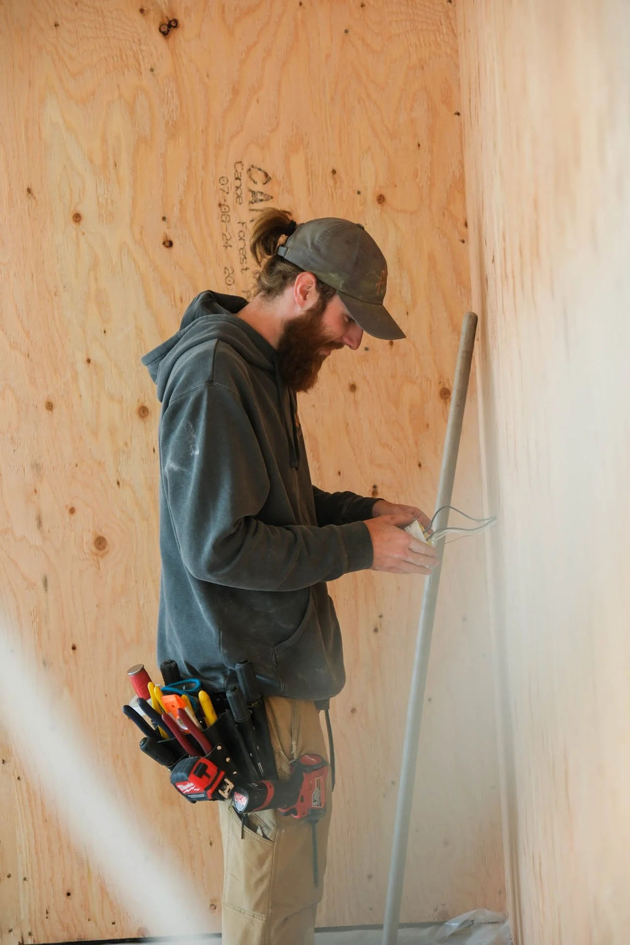 A man with a beard wearing a dark hoodie, beige work pants, and a camouflage cap is working on electrical wiring inside a wooden wall. He has a tool belt with various tools, including screwdrivers and pliers, around his waist. The wall is made of light-colored plywood, and he is standing next to a conduit pipe.