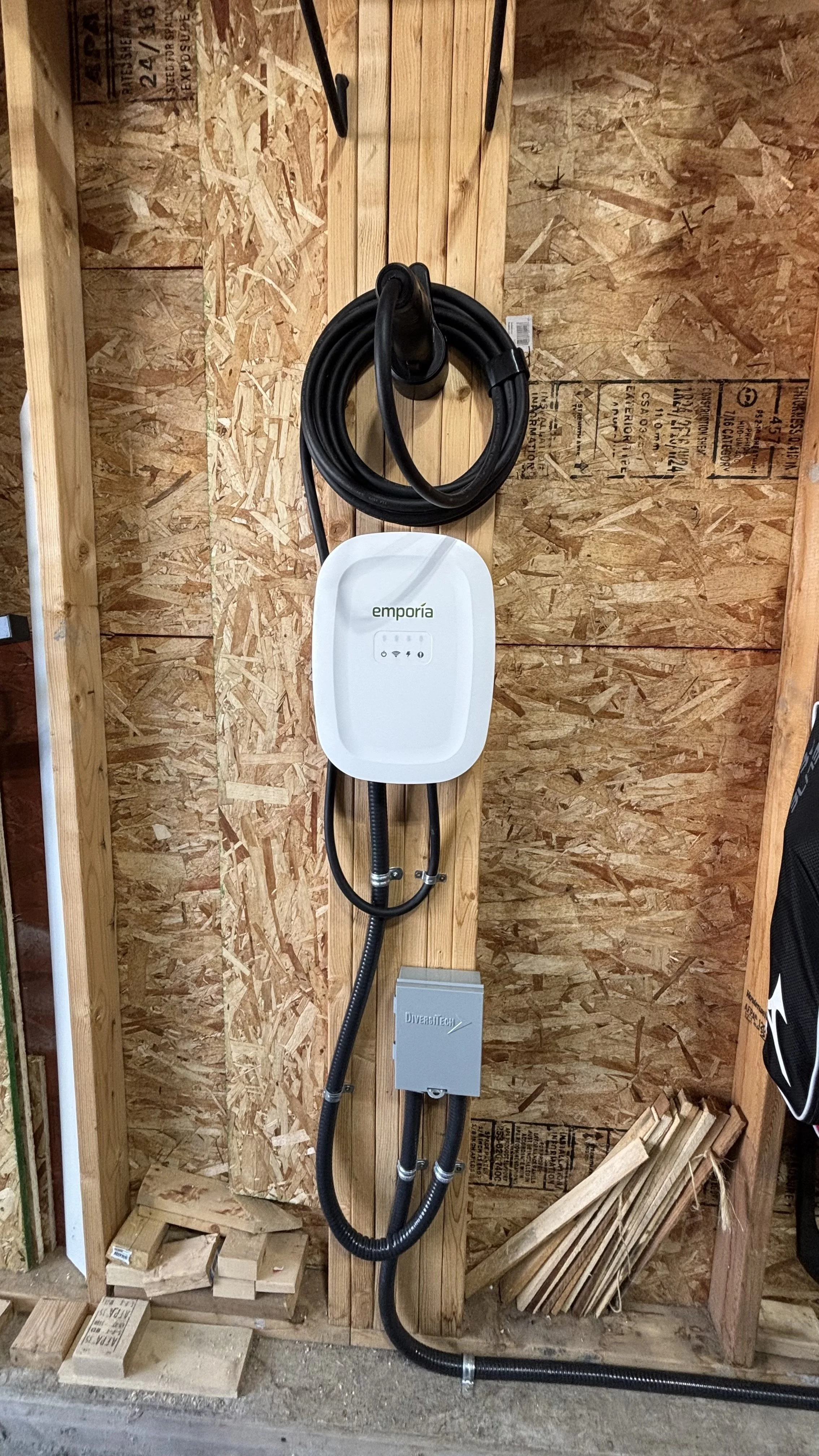 An electric vehicle charging station mounted on an unfinished wooden wall, with black charging cables and a white control box labeled "emporía," in a construction or workshop setting.
