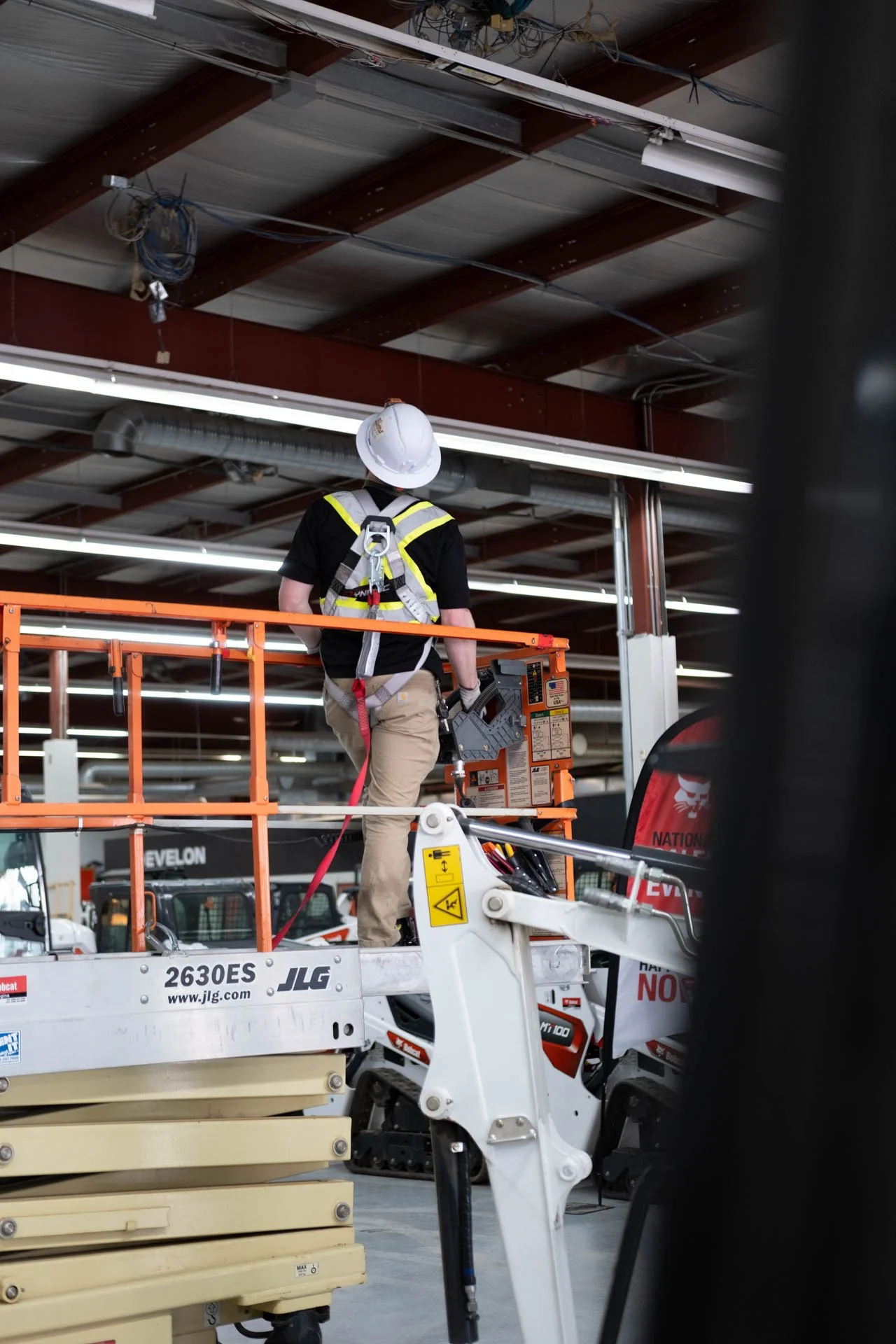 A construction worker on an orange lift, wearing a white helmet and safety harness, working inside a building with exposed ceiling beams, pipes, and fluorescent lighting.