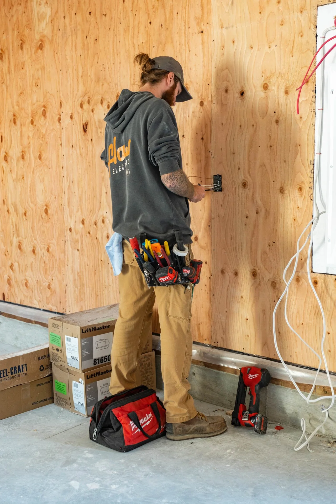 A construction worker standing by a wooden wall, working with electrical wiring, with tools and boxes around him.