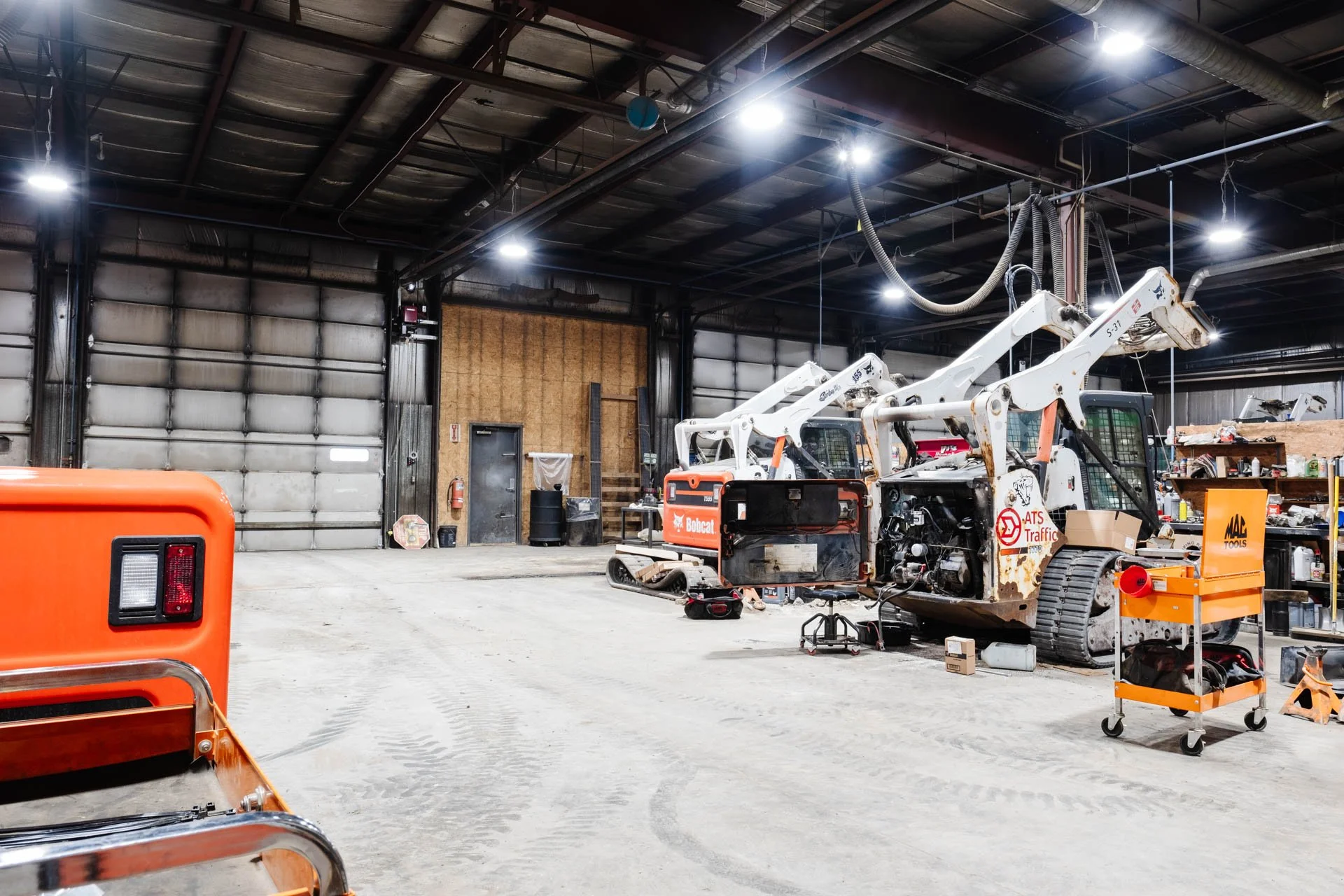 Industrial workshop with construction equipment, tools, and machinery, including a Bobcat loader, in a spacious indoor setting with high ceilings and overhead lighting.