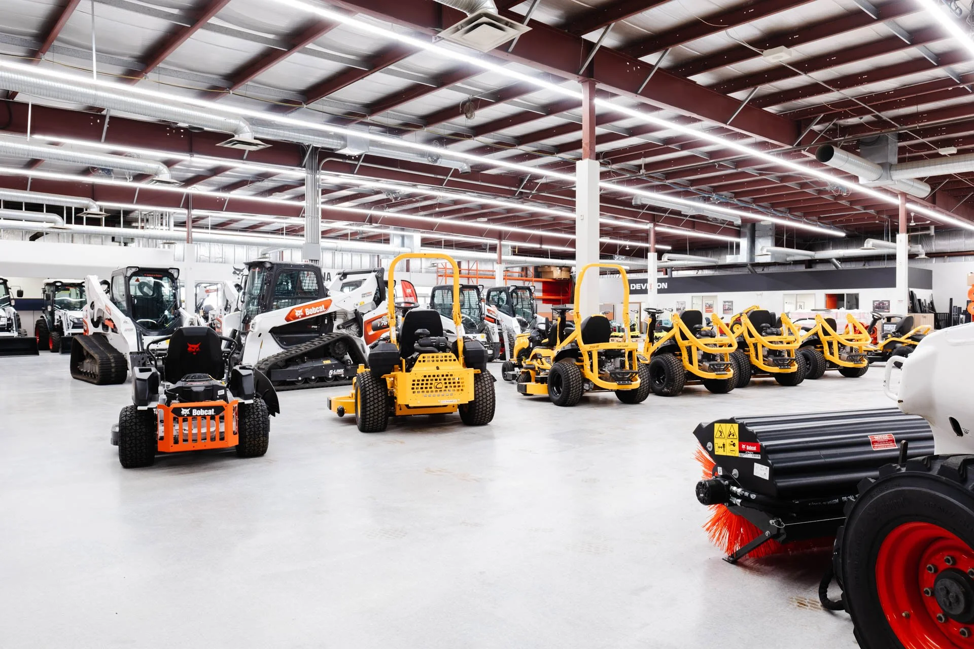 Interior of a warehouse or showroom with various construction and landscaping equipment, including small bulldozers, ride-on mowers, and other machinery, arranged in an organized manner under a metal ceiling with visible ductwork and lighting.