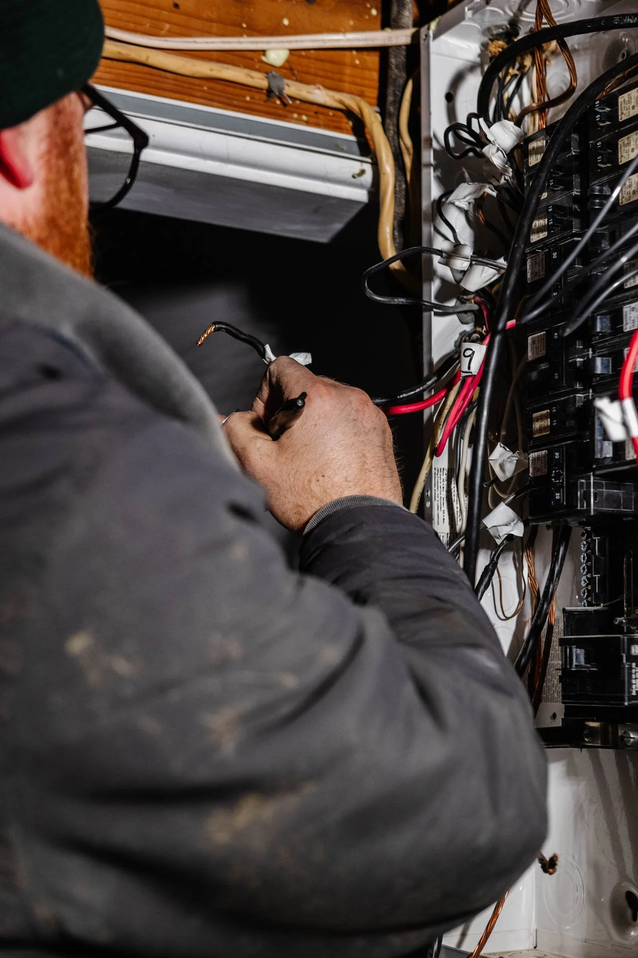 A technician with a beard, wearing glasses, black cap, and gray work clothes, working on electrical wiring inside an electrical panel.