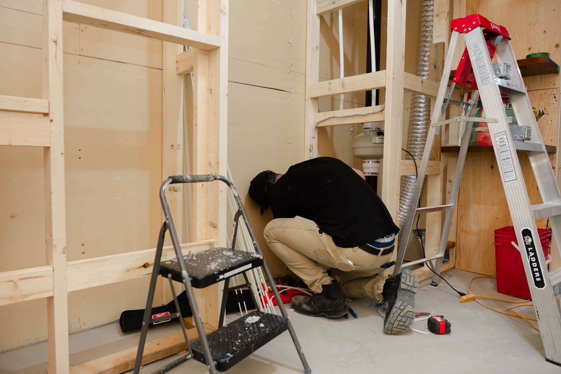 A person working on wiring or electrical work inside a partially constructed room with exposed wooden framing, a metal step ladder, and various tools.