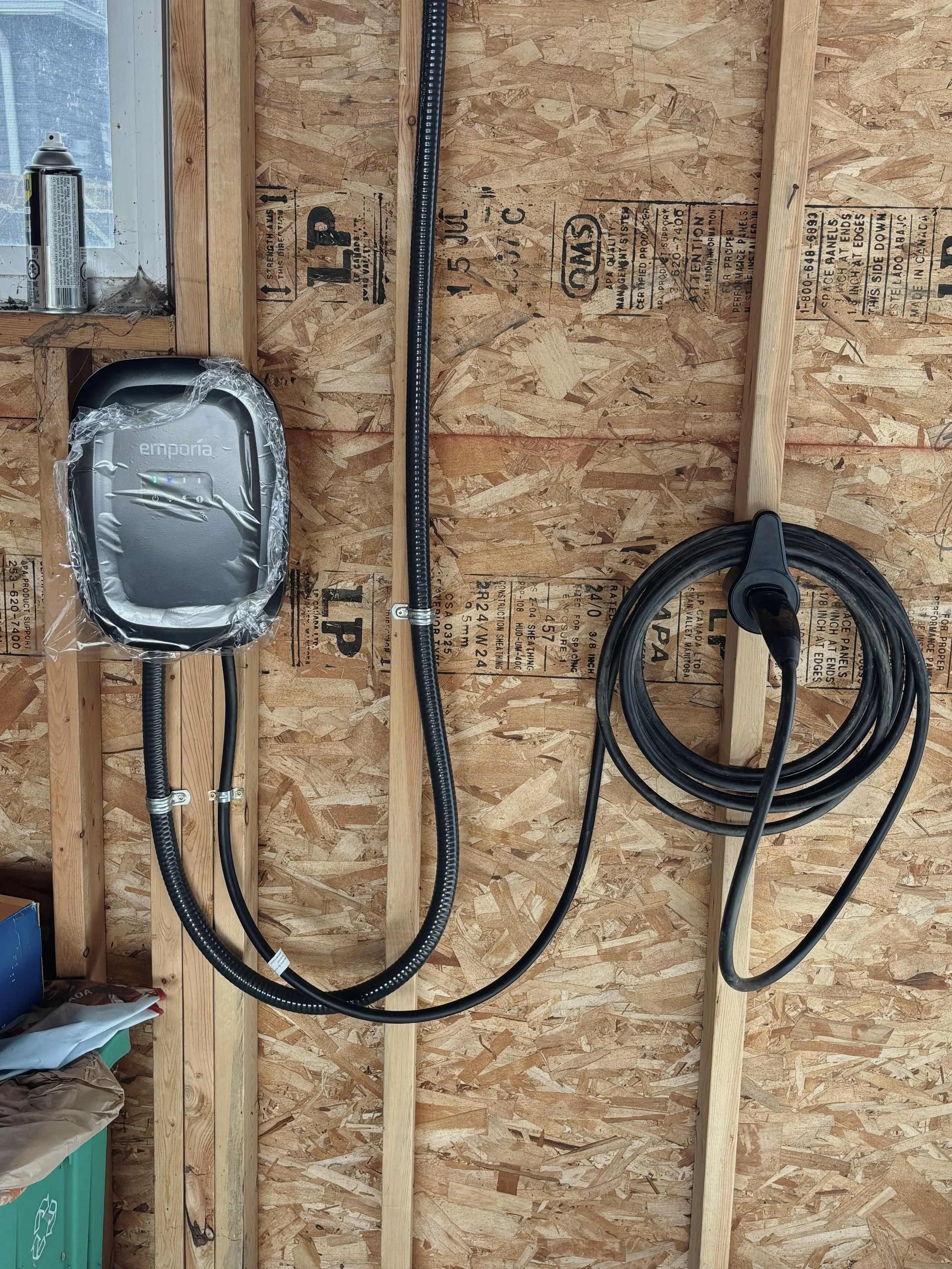 An electric vehicle charging station mounted on plywood wall with a connected black charging cable, a white control unit labeled 'Emporia,' and a gray box labeled 'Diverse Tech' at the bottom, surrounded by construction materials.