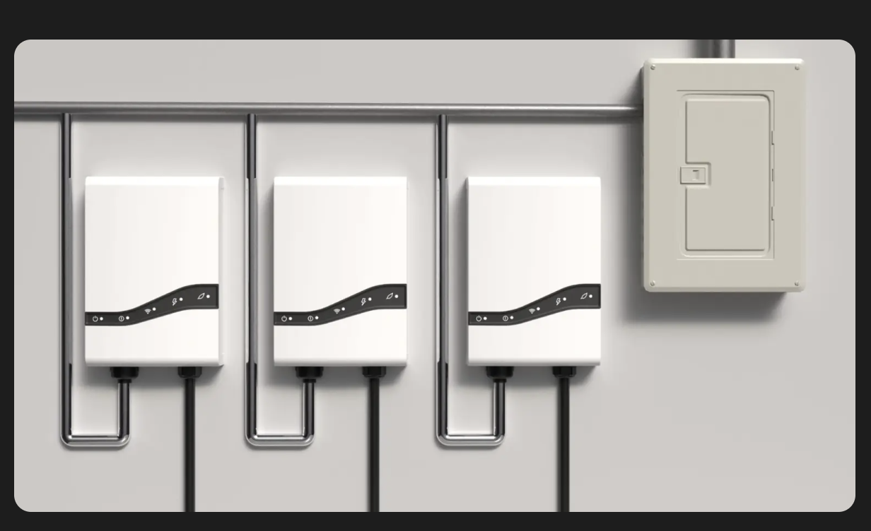 Three white electrical inverters mounted on a gray wall with black wires connected to each, and a gray electrical panel box to the right.
