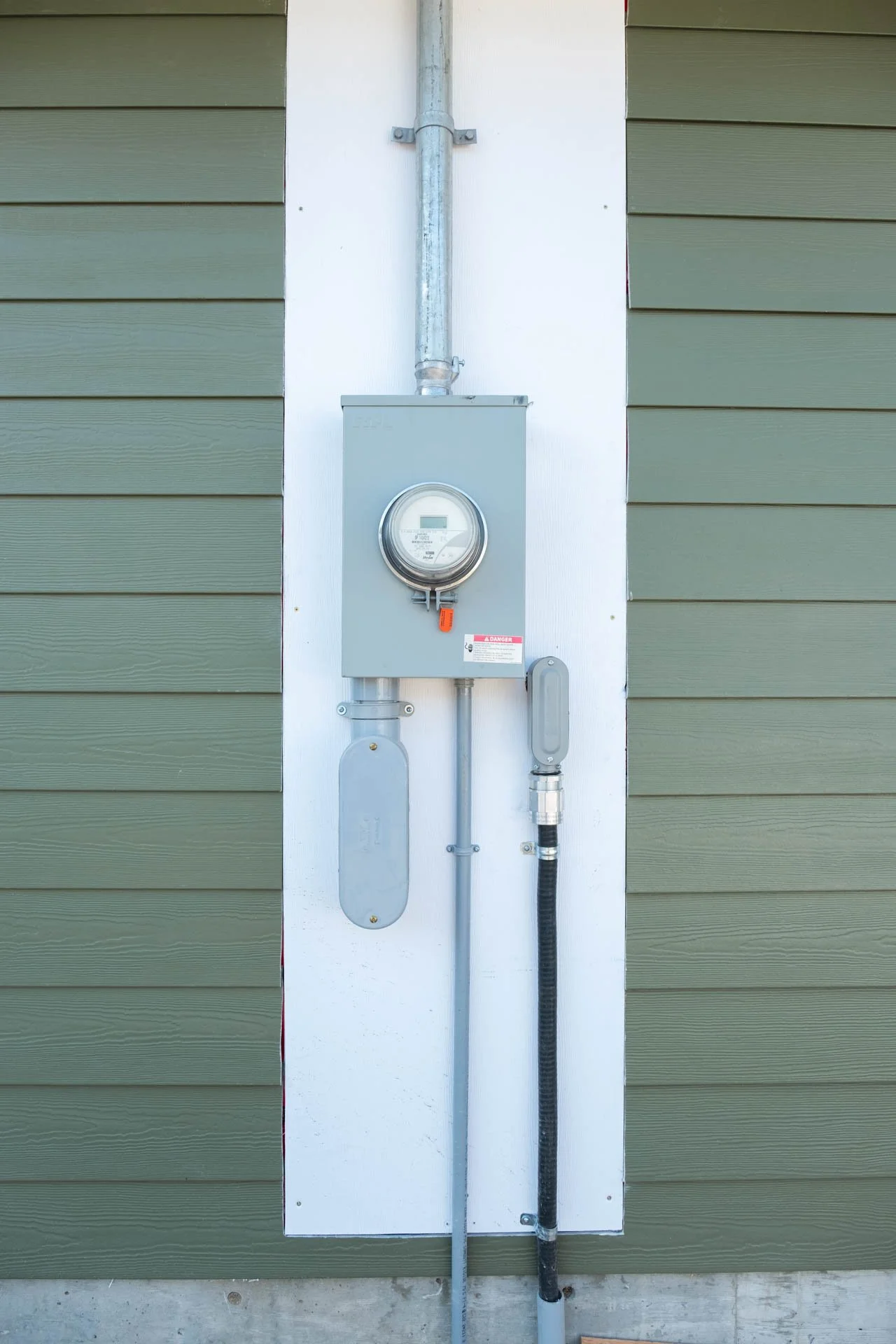 Electric meter mounted on a utility box on the exterior wall of a building with green siding.