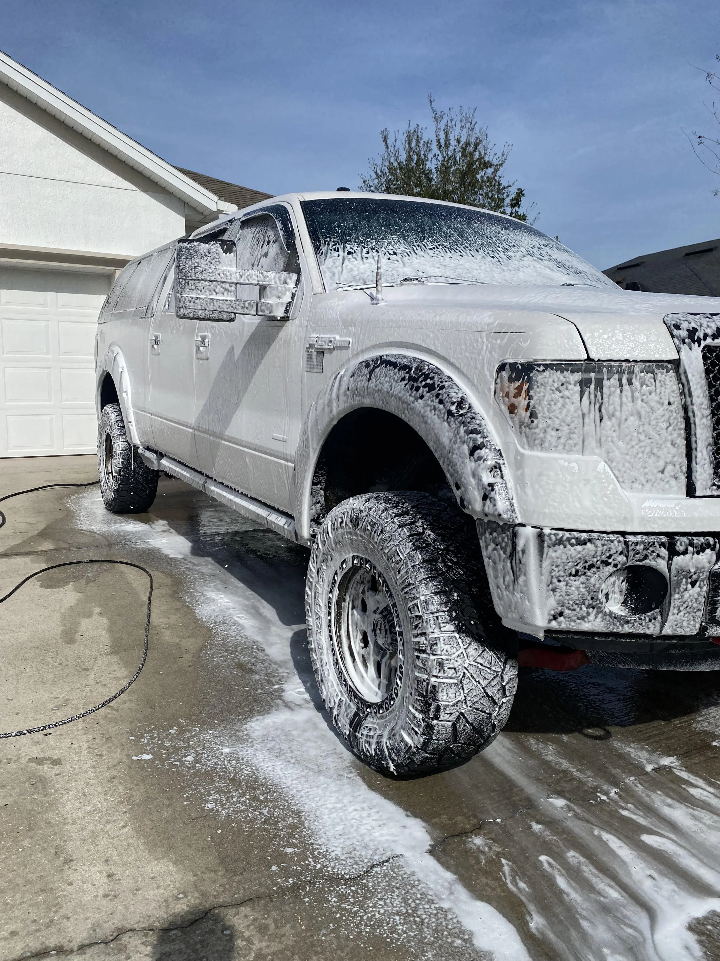 A large SUV covered in soap suds on a driveway, undergoing a car wash in front of a residential garage.