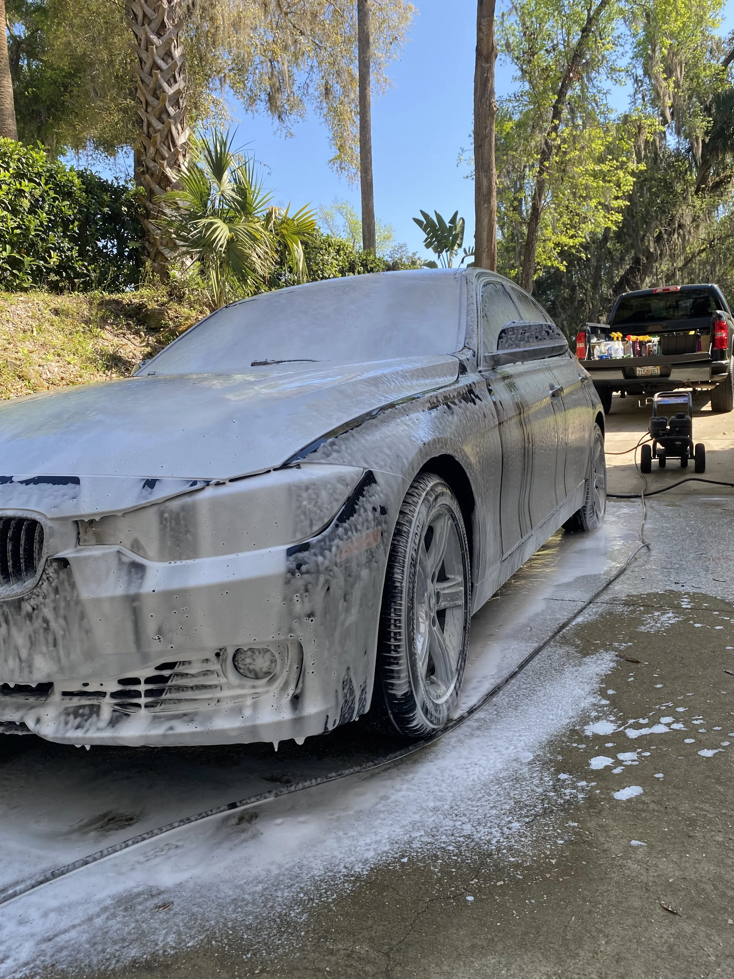 Car covered in soap foam during a wash, surrounded by trees and a parked truck in the background.