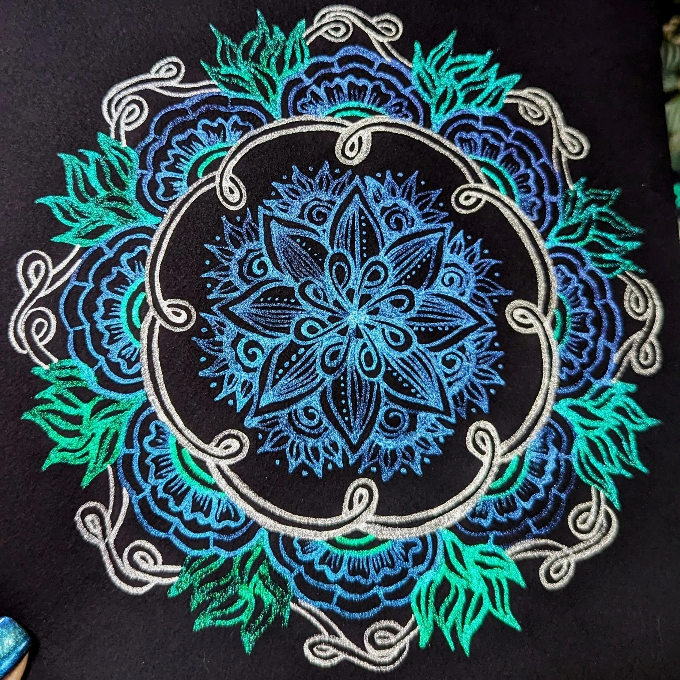 Intricate blue and green mandala design on black background with floral and swirl patterns.