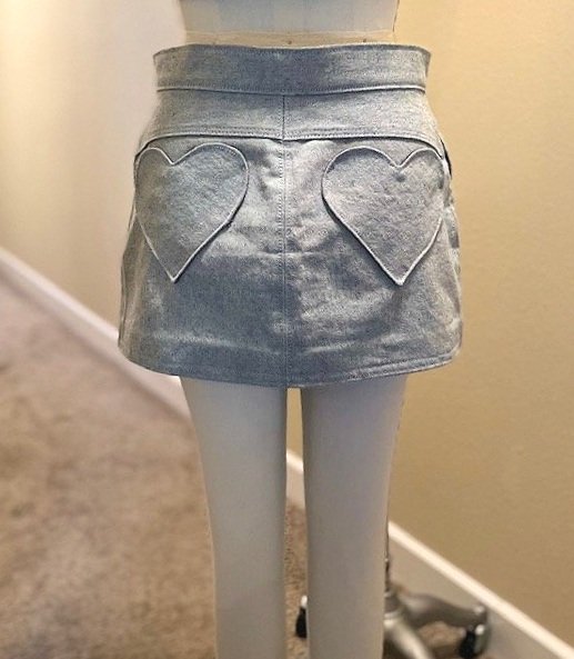 Back of skirt. Denim mini skirt in gray. Heart shaped pockets with a front fly and zipper. White button.