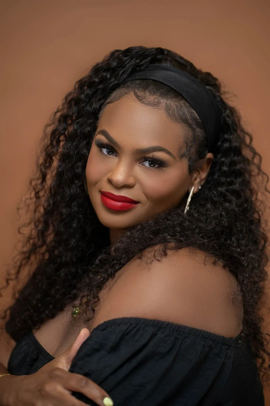 Smiling woman with curly hair, red lipstick, and off-the-shoulder black top against a brown background.