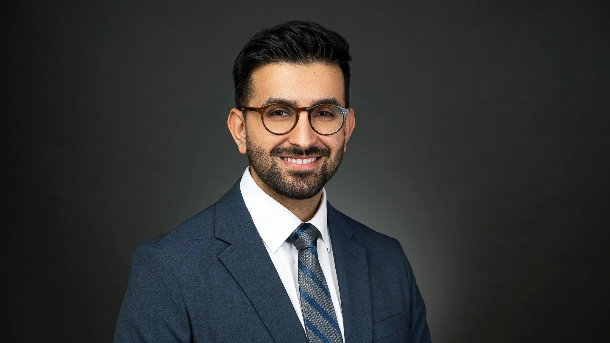Smiling man in a suit and tie with glasses against a dark background.