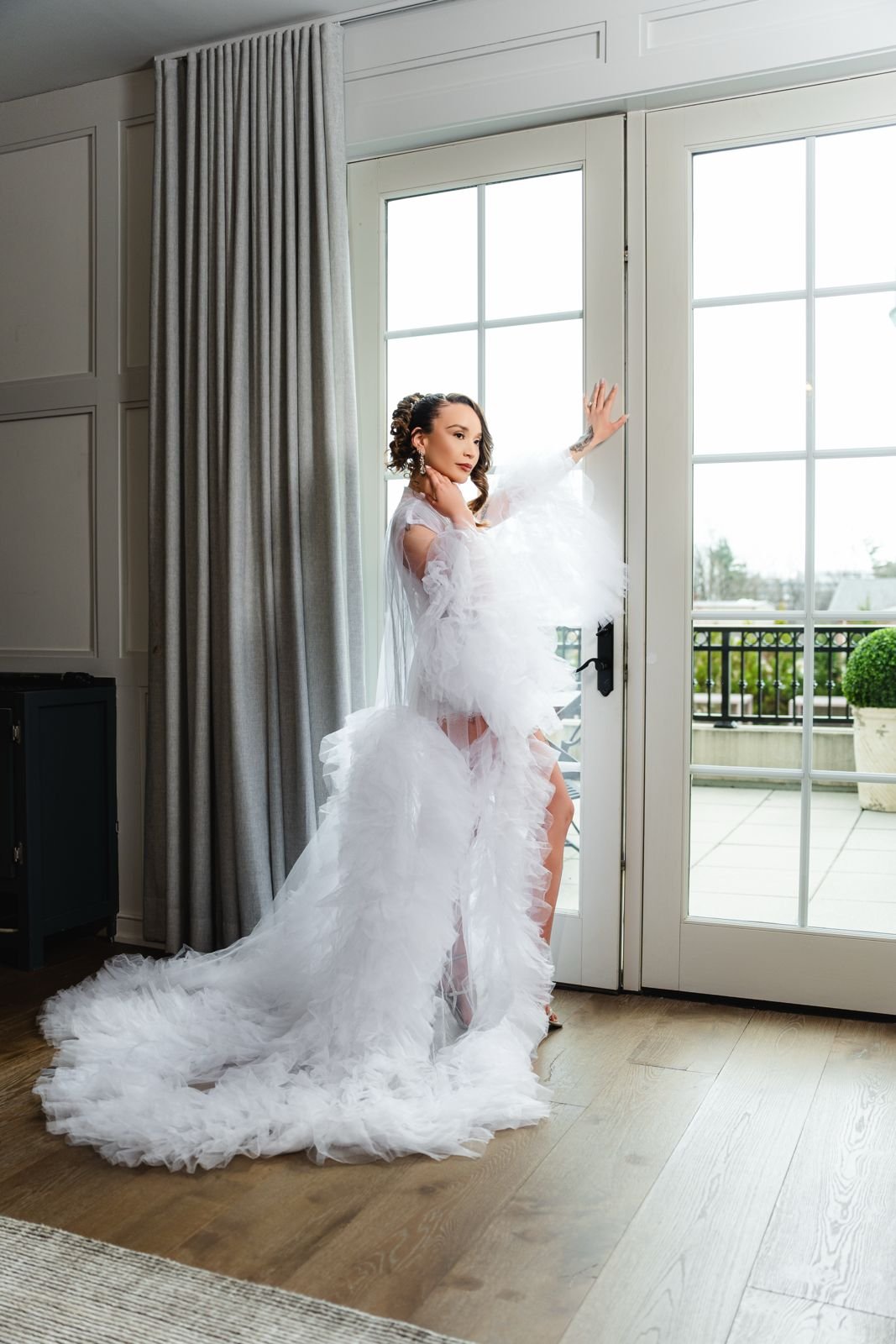 Person in a flowing white gown standing by glass doors, looking outside.