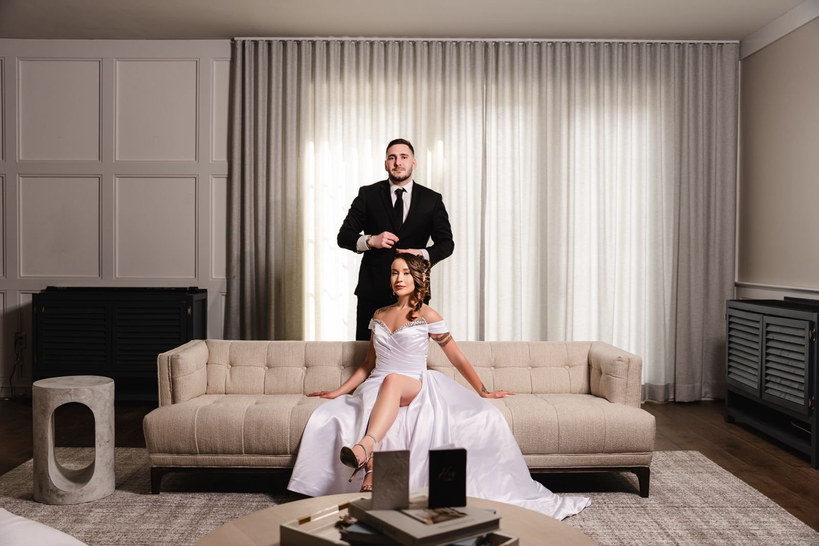 A man in a black suit and a woman in a white gown seated on a beige sofa in a sophisticated living room with curtains.