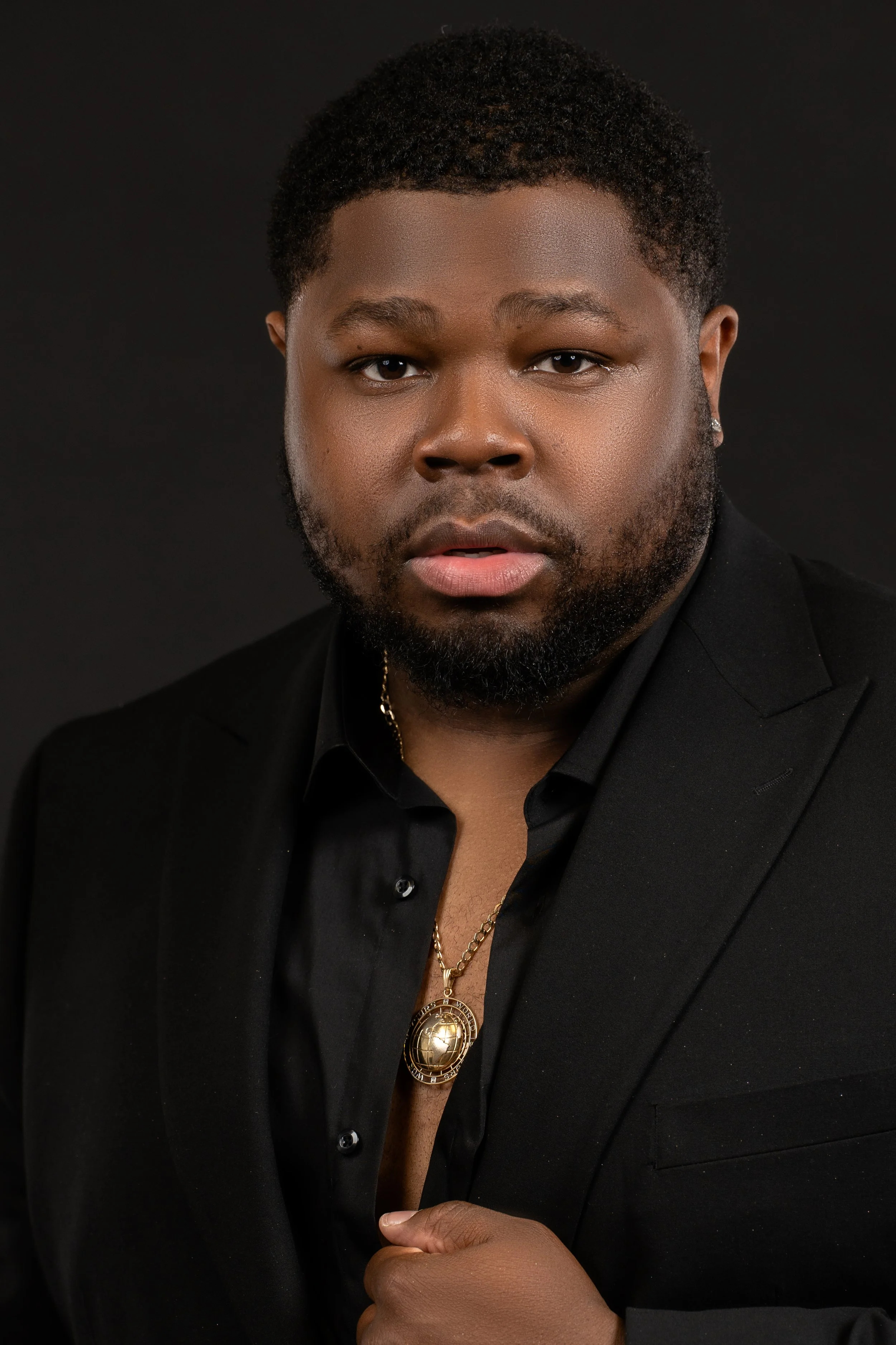 Man in black suit and shirt with gold necklace against dark background.
