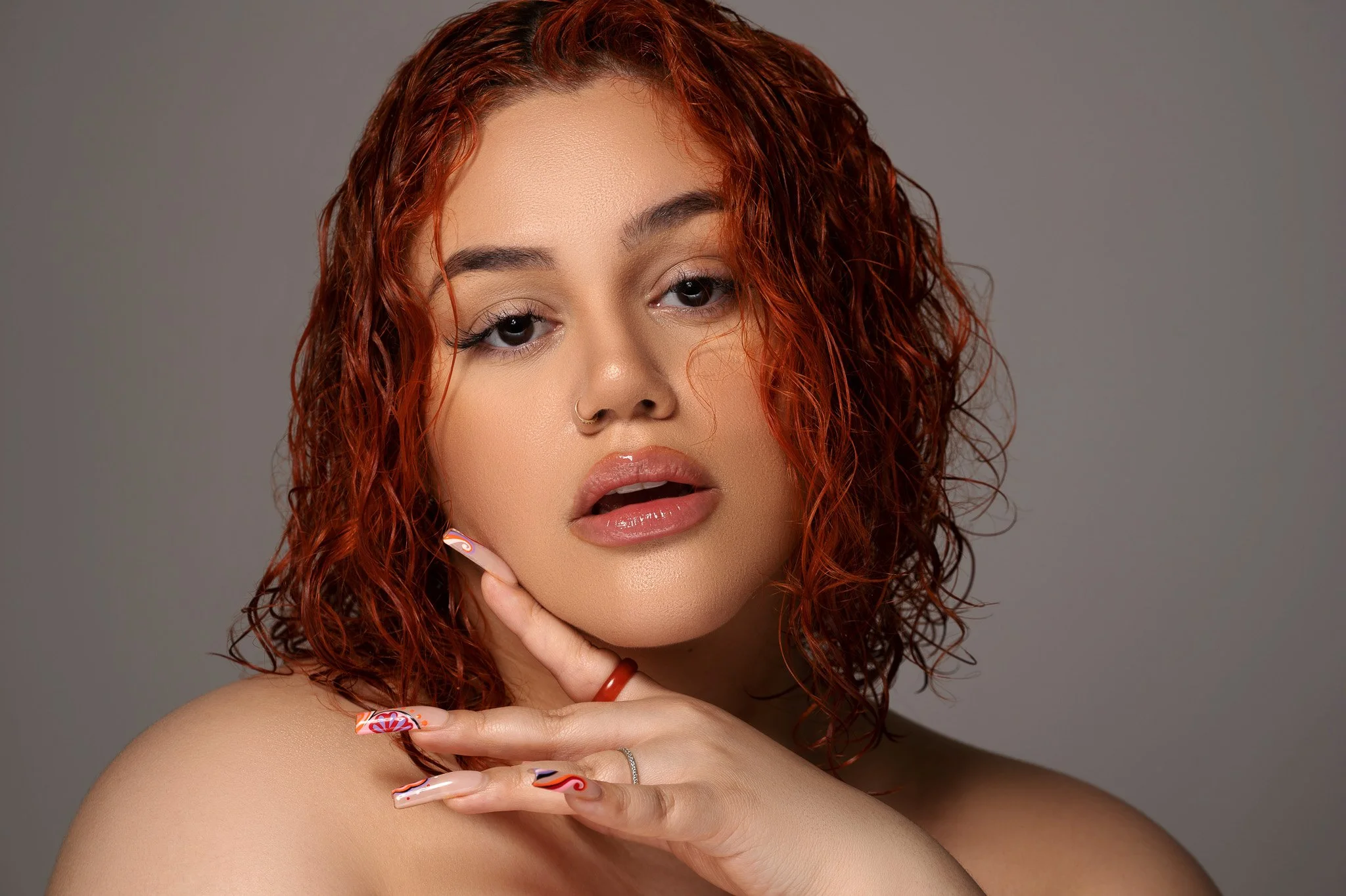 A person with red, curly hair, wearing a nose ring, glossy lipstick, and nail art, posing with their hand under their chin against a neutral background.