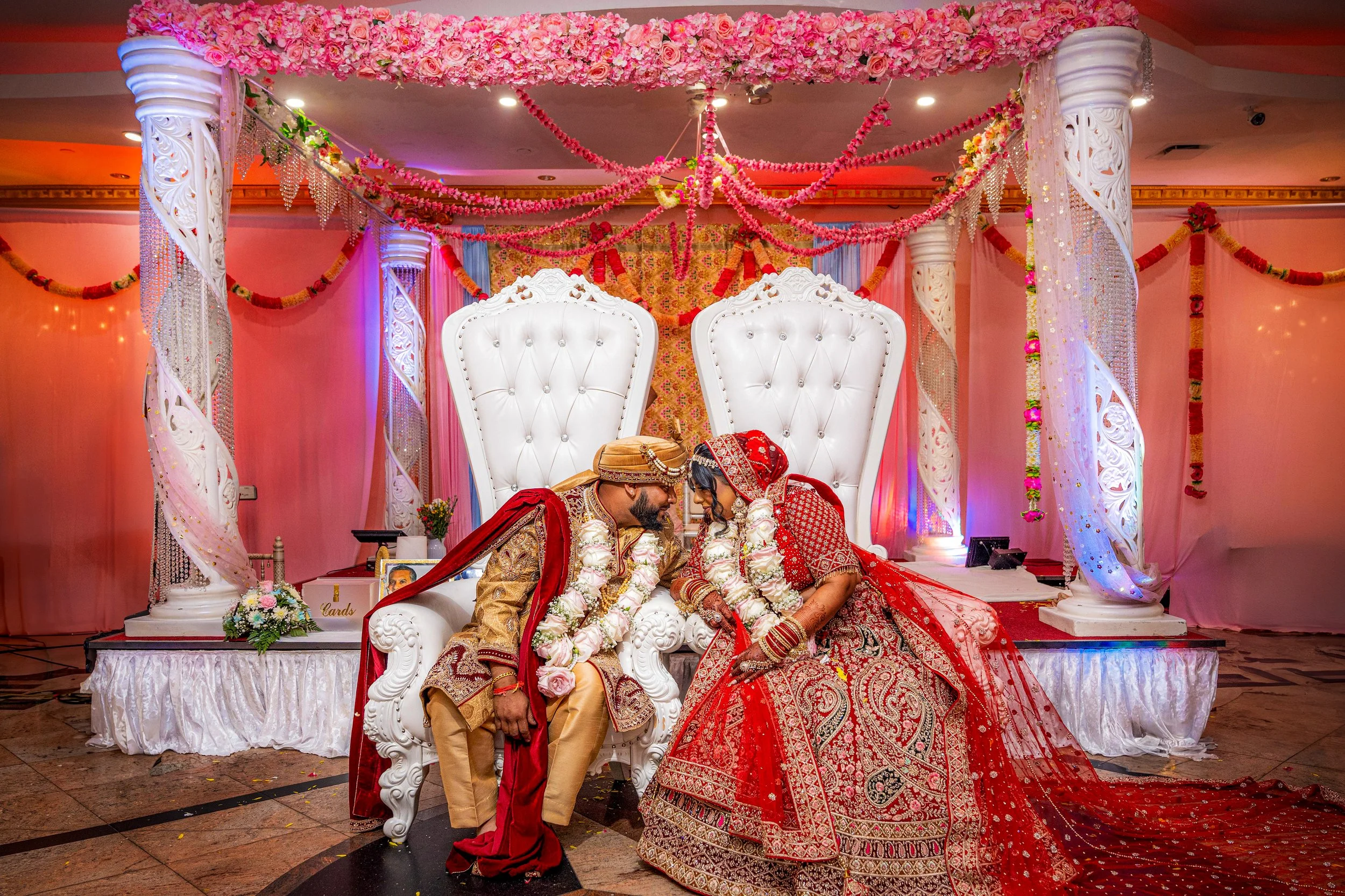 Bride and groom in traditional attire, surrounded by floral decorations, seated on ornate white chairs, partaking in a wedding ceremony.
