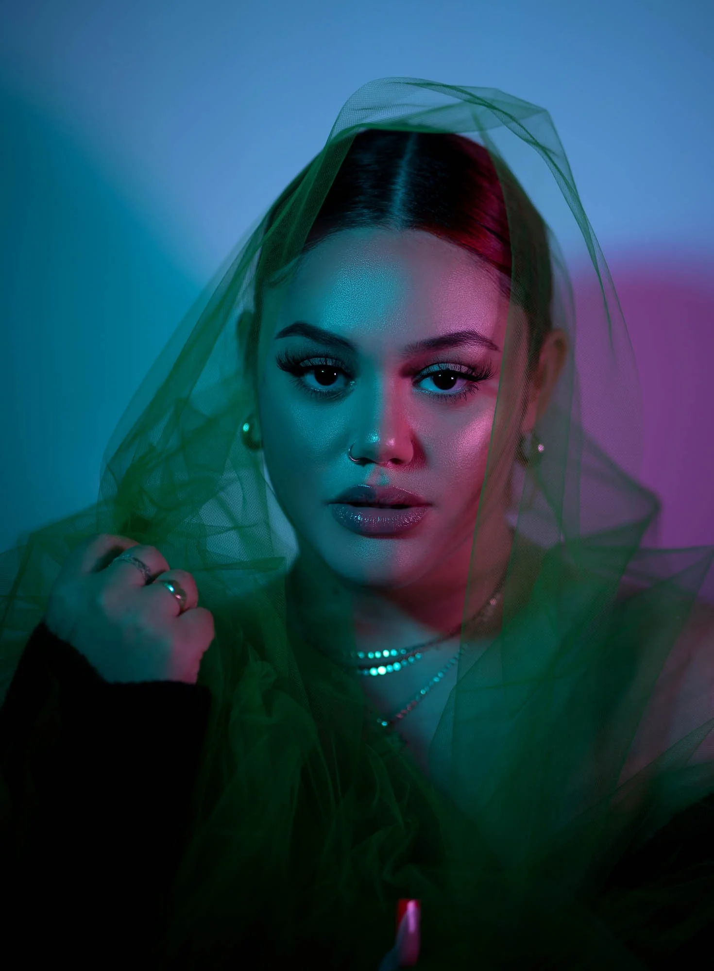 A person with makeup and a nose ring is partially covered by a green veil, under blue and pink lighting.