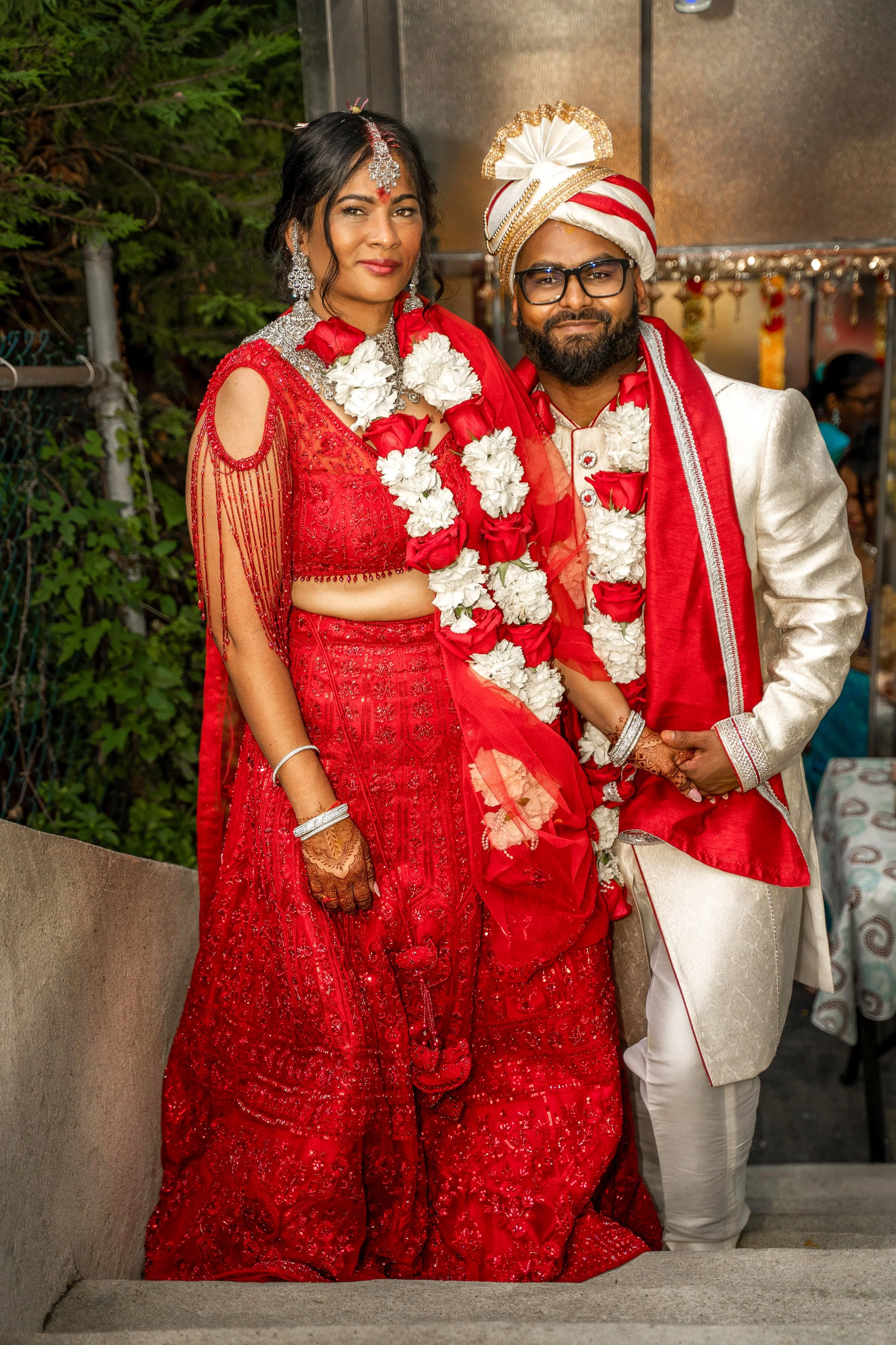 A couple in traditional Indian wedding attire, with the woman wearing a red lehenga and the man in a white sherwani and red turban, both adorned with flower garlands.