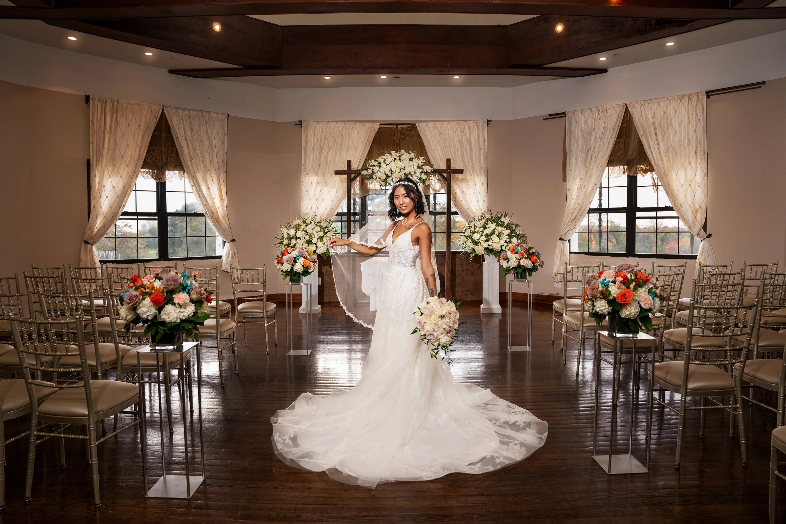 Bride in white wedding dress holding bouquet, standing in decorated indoor venue with flowers and chairs.