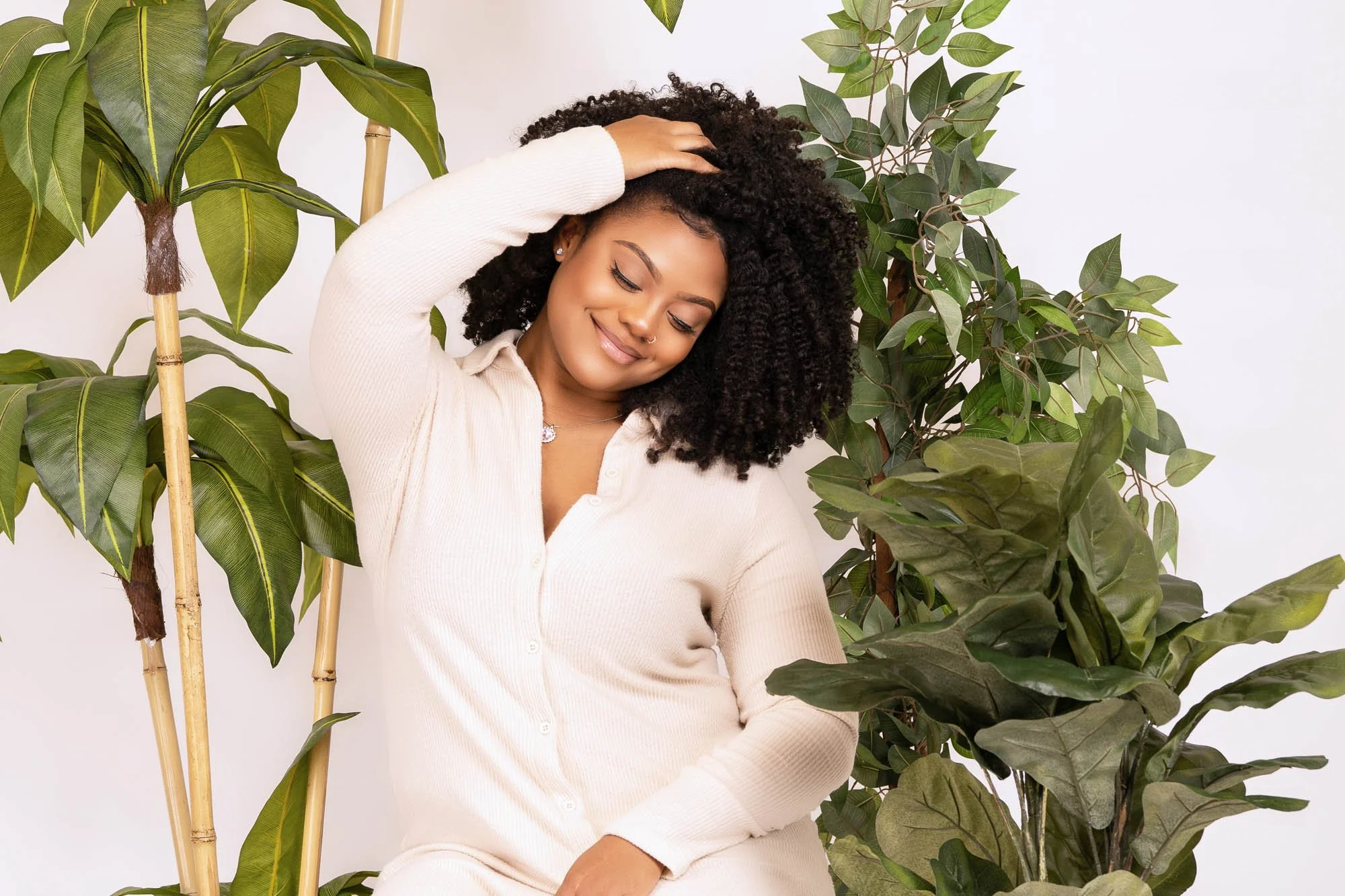 Smiling woman with curly hair wearing a white cardigan, sitting among green plants.