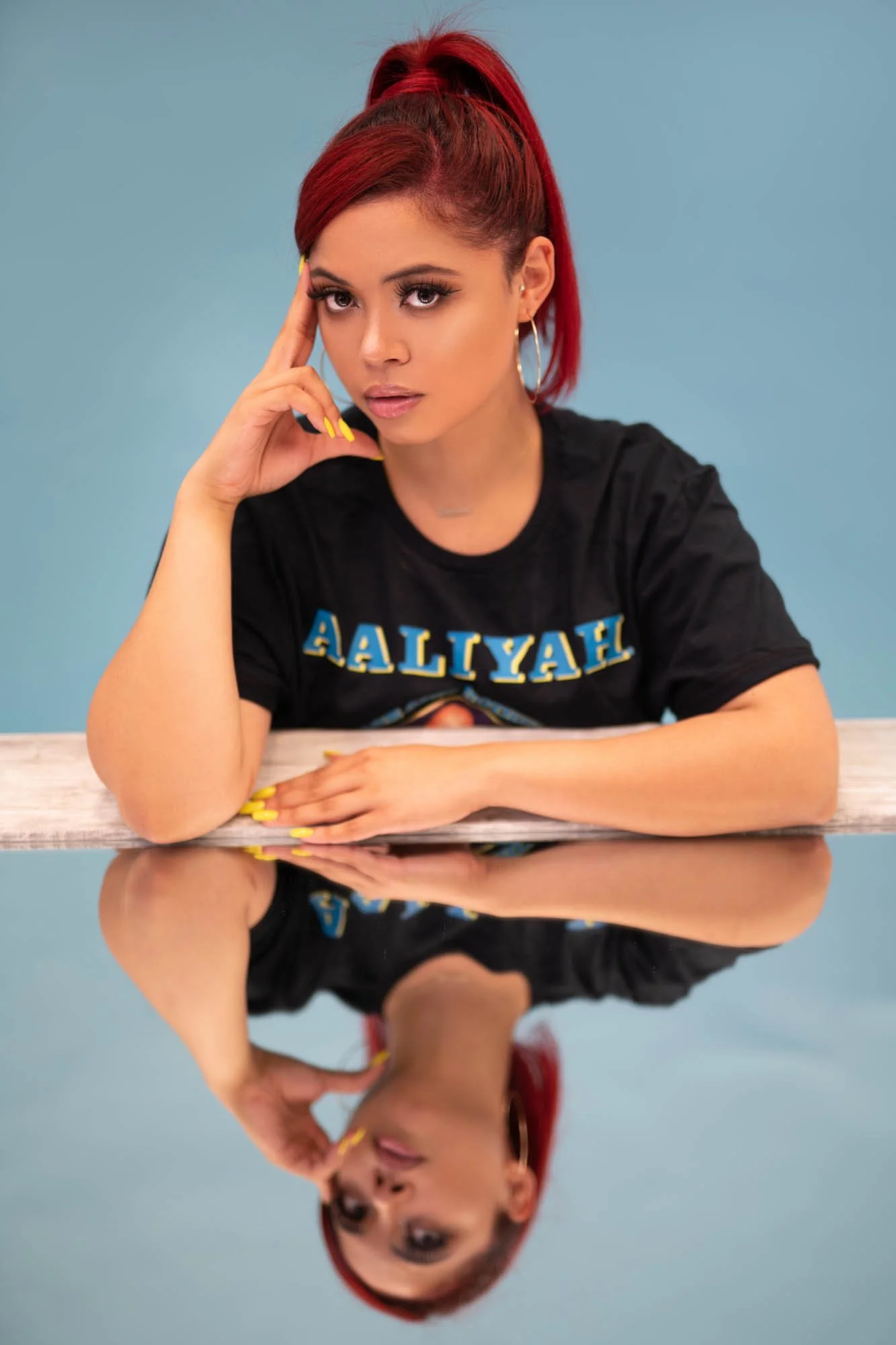 Woman with red hair and hoop earrings, wearing a black Aaliyah t-shirt, resting her chin on her hand with mirror reflection below, light blue background.