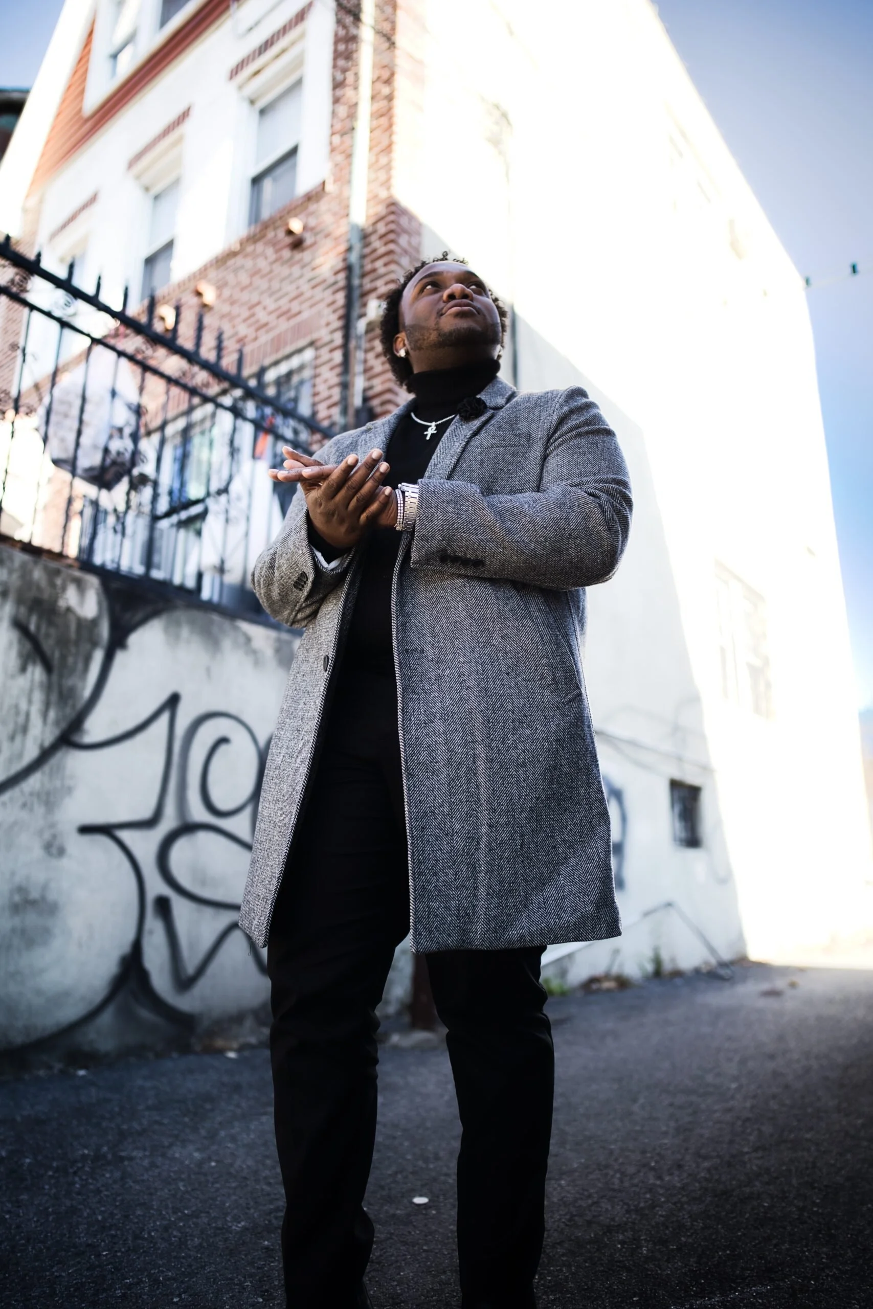 Person in gray coat and black pants standing on urban street with graffiti and buildings, looking up.