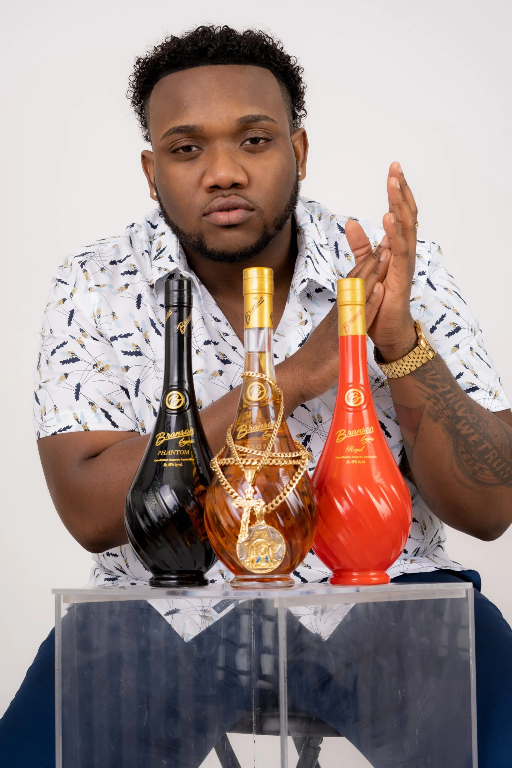 A person with short curly hair wearing a printed shirt and sitting behind three bottles of Branson Cognac in black, brown, and red on a clear table. The middle bottle is adorned with a gold chain and medallion.