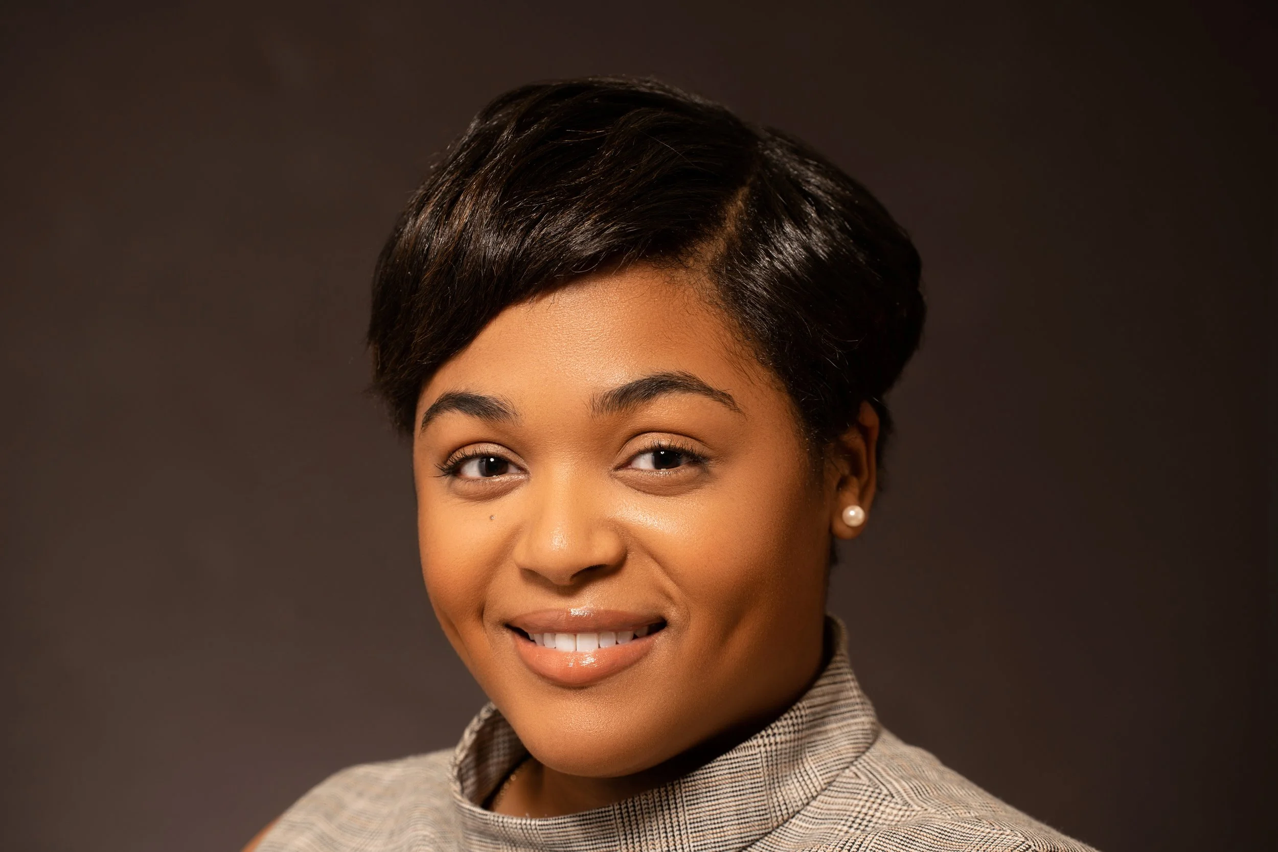Portrait of a smiling person with short dark hair, wearing a checkered top and pearl earrings, against a dark background.