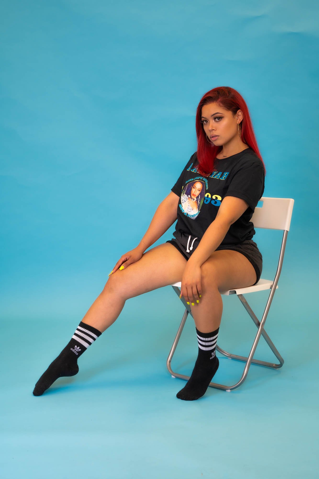 A person with red hair wearing a black graphic t-shirt and shorts sits on a white chair against a blue background. They have black striped socks and black socks. The t-shirt features a printed portrait.