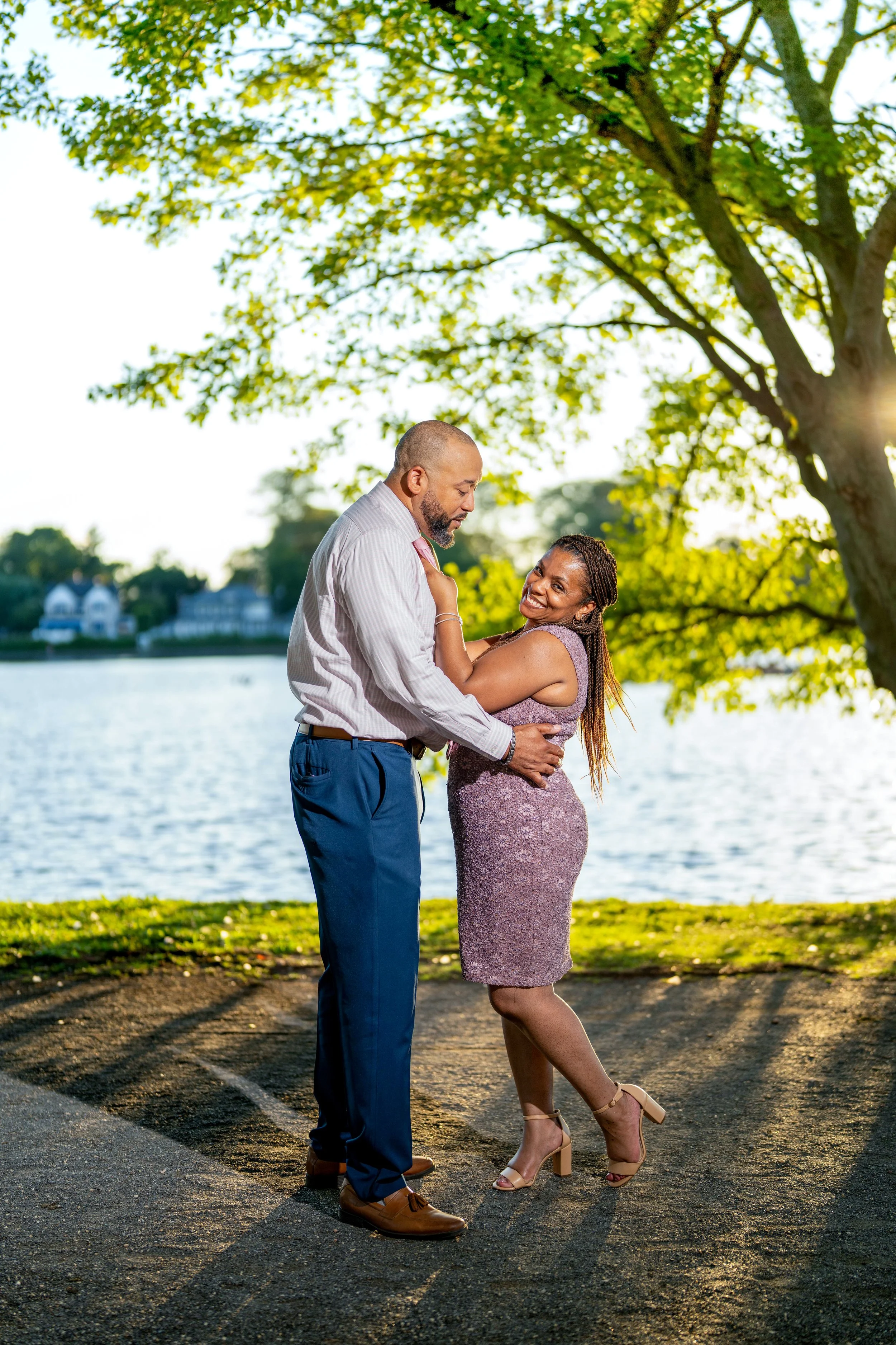 A couple embracing and smiling by a lake, surrounded by trees, with sunlight filtering through the leaves.