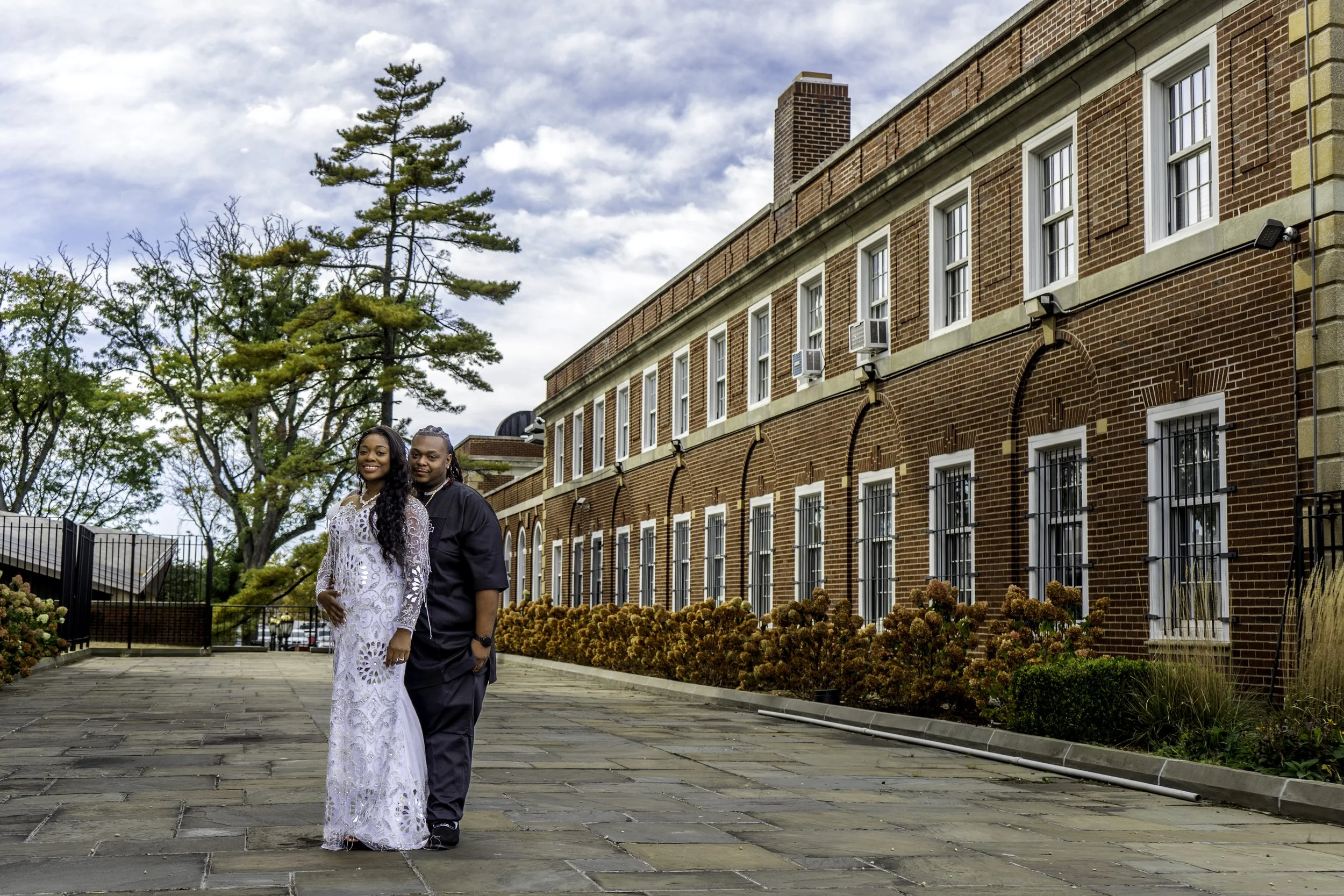 Couple posing outdoors with a brick building and trees in the background.