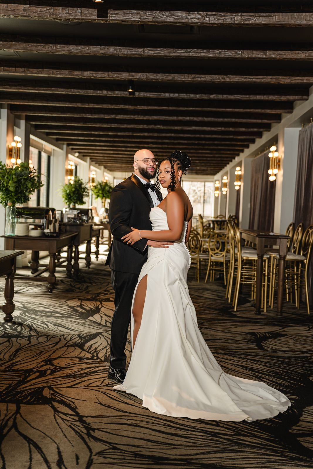 A couple posing in a decorated venue, the woman wearing a white wedding dress and the man in a black suit. The space has wooden beams, decorative lighting, and greenery.