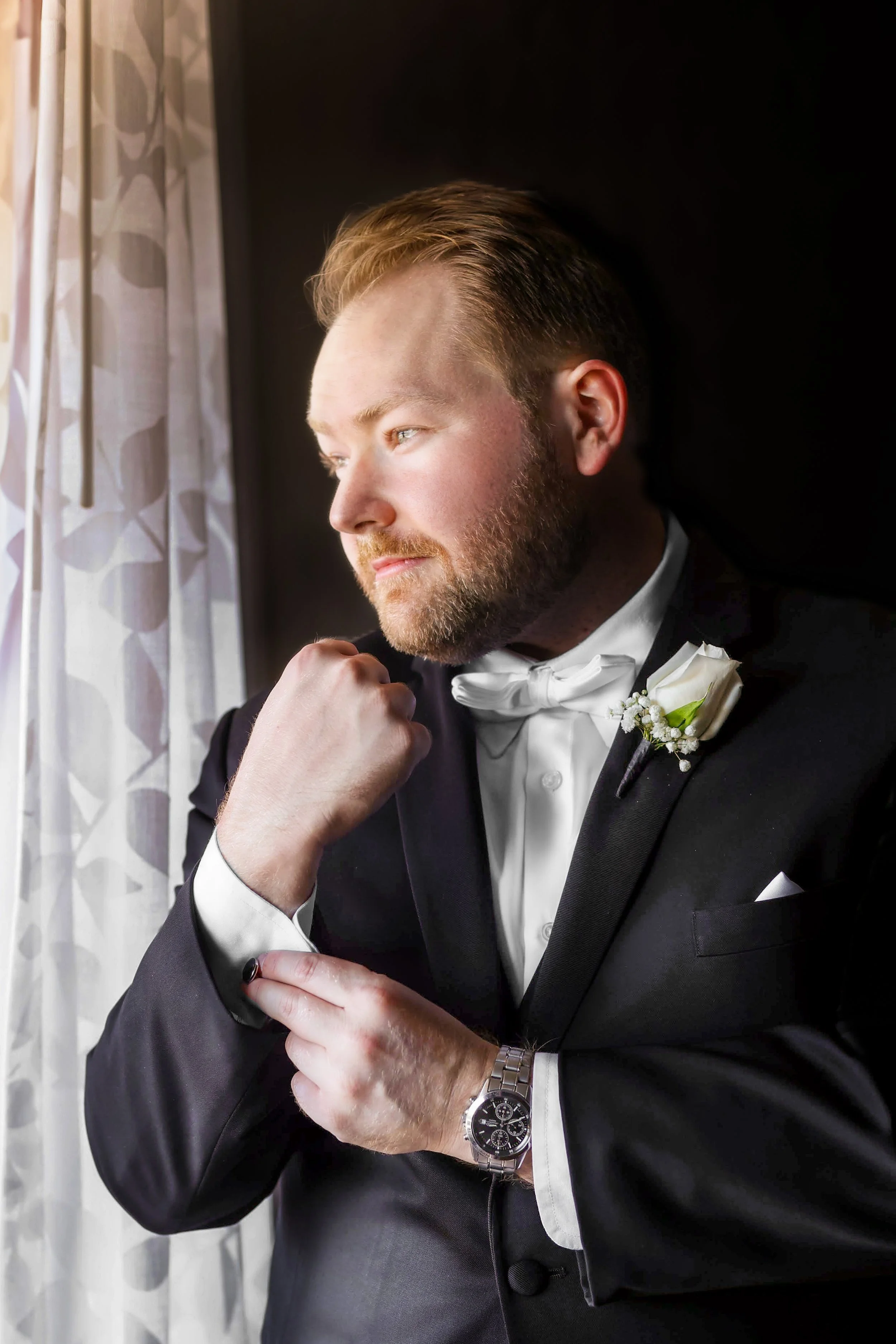 Man in a tuxedo adjusting cufflink, wearing a bow tie and boutonniere.