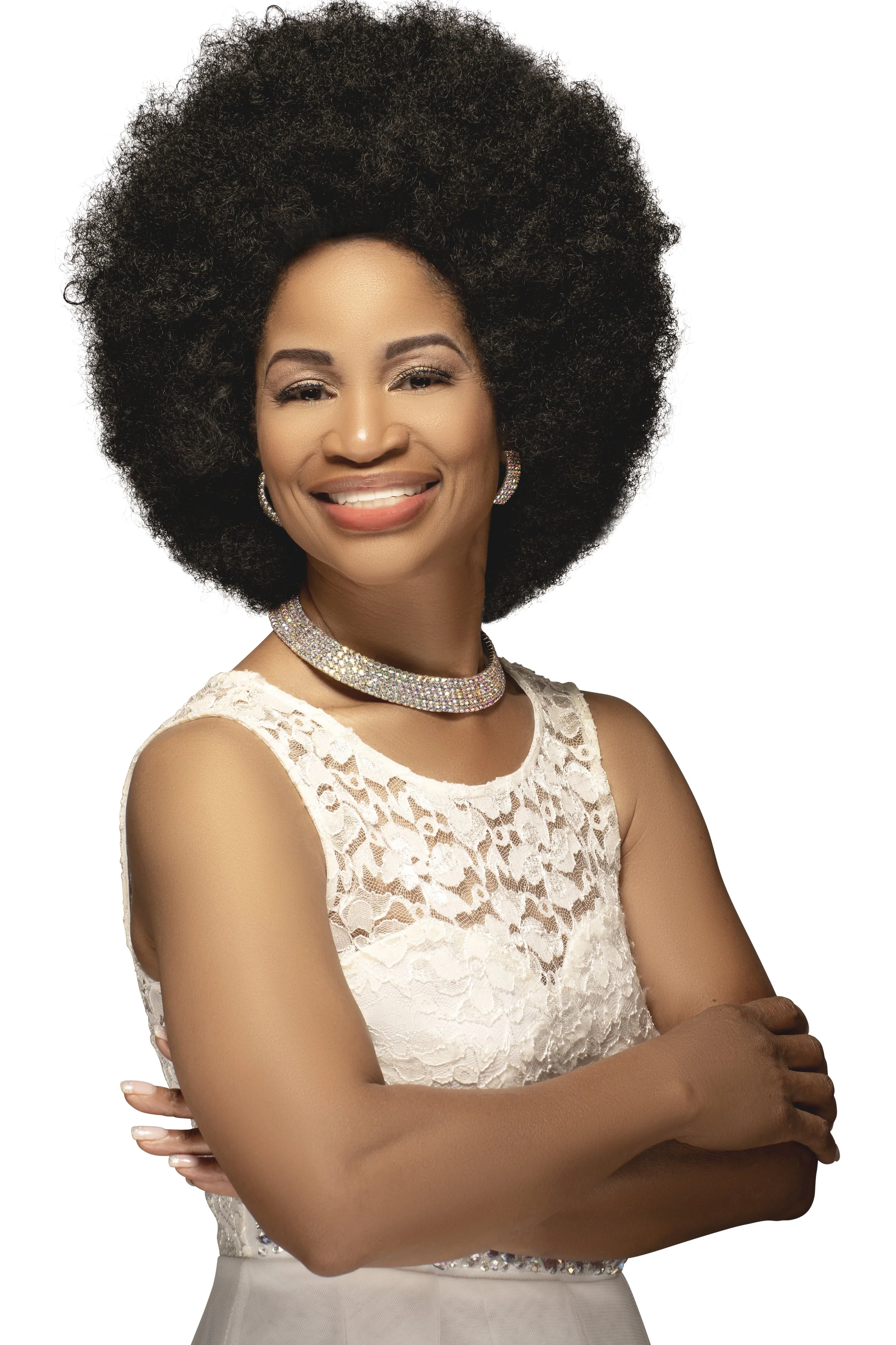 Smiling woman with an afro hairstyle, wearing a white lace dress and jewelry, arms crossed.