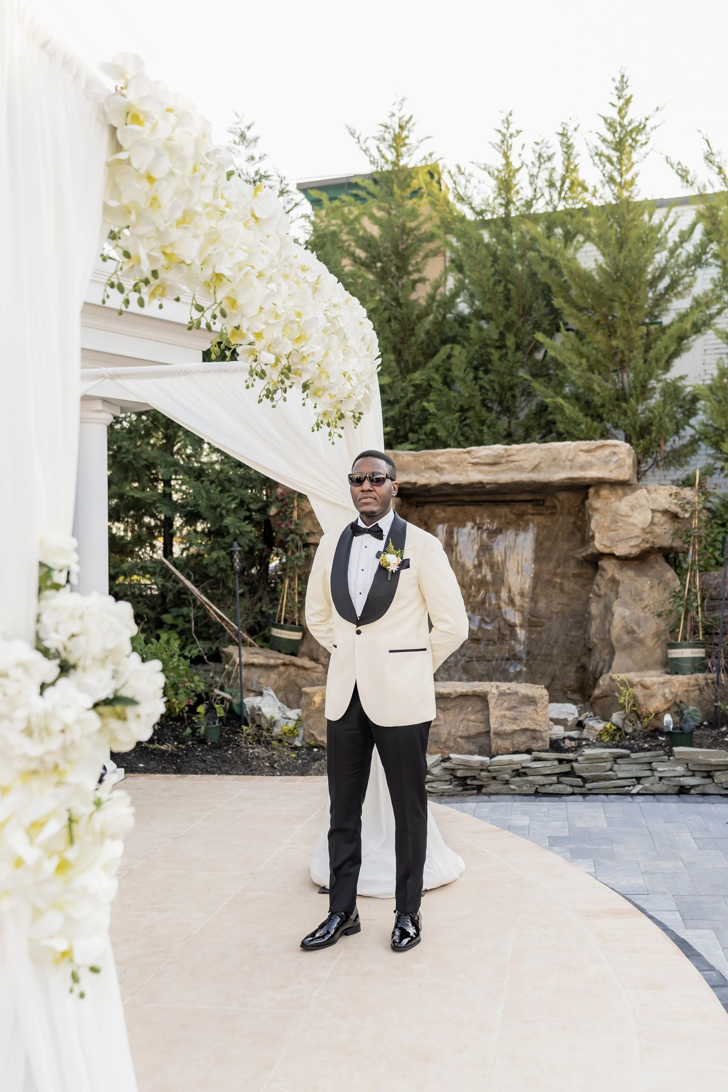 Person in a white and black tuxedo standing outdoors under a flower-adorned archway, surrounded by greenery and a stone waterfall.