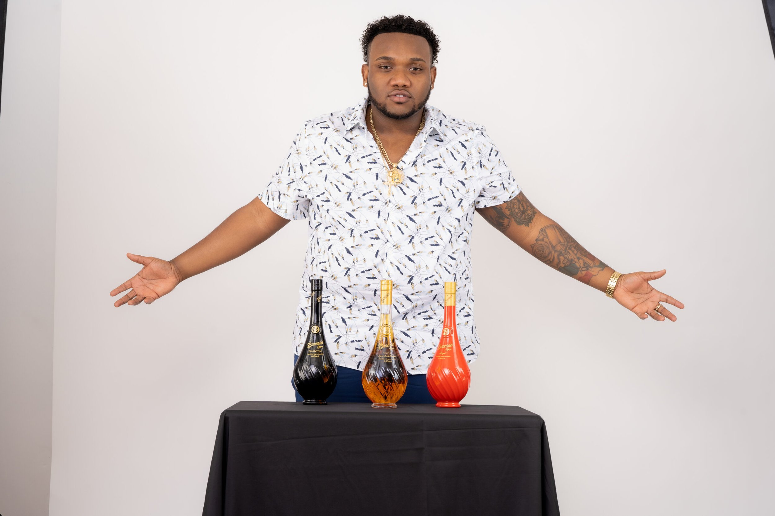 Person presenting three colorful liquor bottles on a table with a black tablecloth, wearing a patterned shirt and gold jewelry.