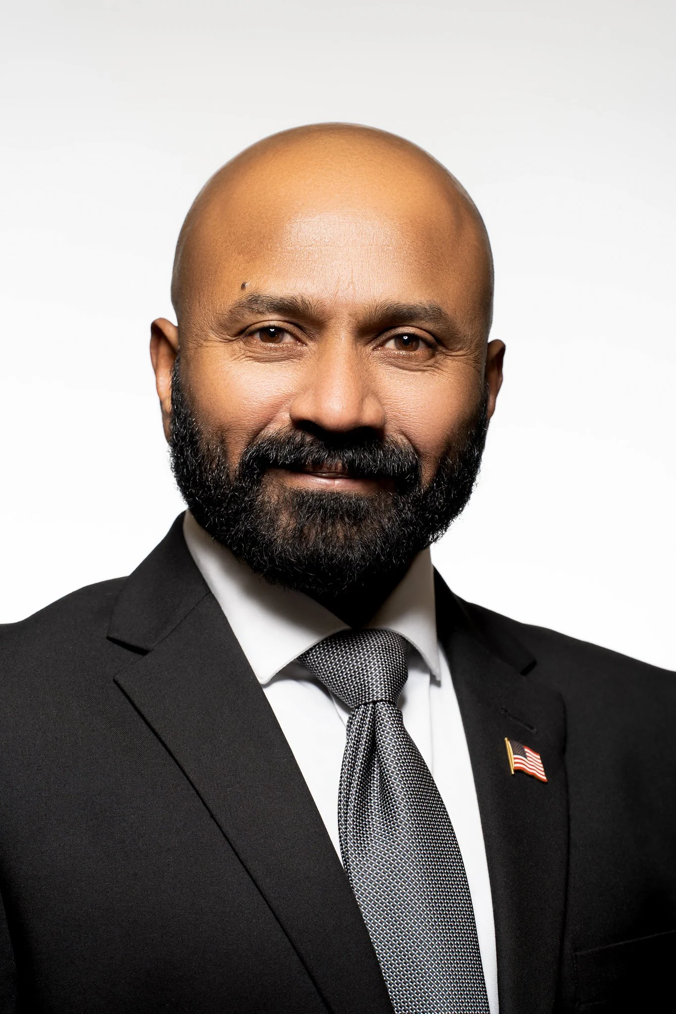 Portrait of a bald man with a beard wearing a suit and tie, featuring an American flag lapel pin.