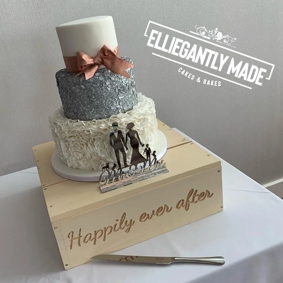 A three-tier wedding cake with white frosting, a silver textured middle layer, and a white top layer with a peach-colored ribbon bow. The cake sits on a white base on a wooden crate with the words "Happily ever after" engraved. There is a cake topper of a bride and groom holding hands with a small child. The backdrop features a sign reading "Elegantly Made Cakes & Bakes."
